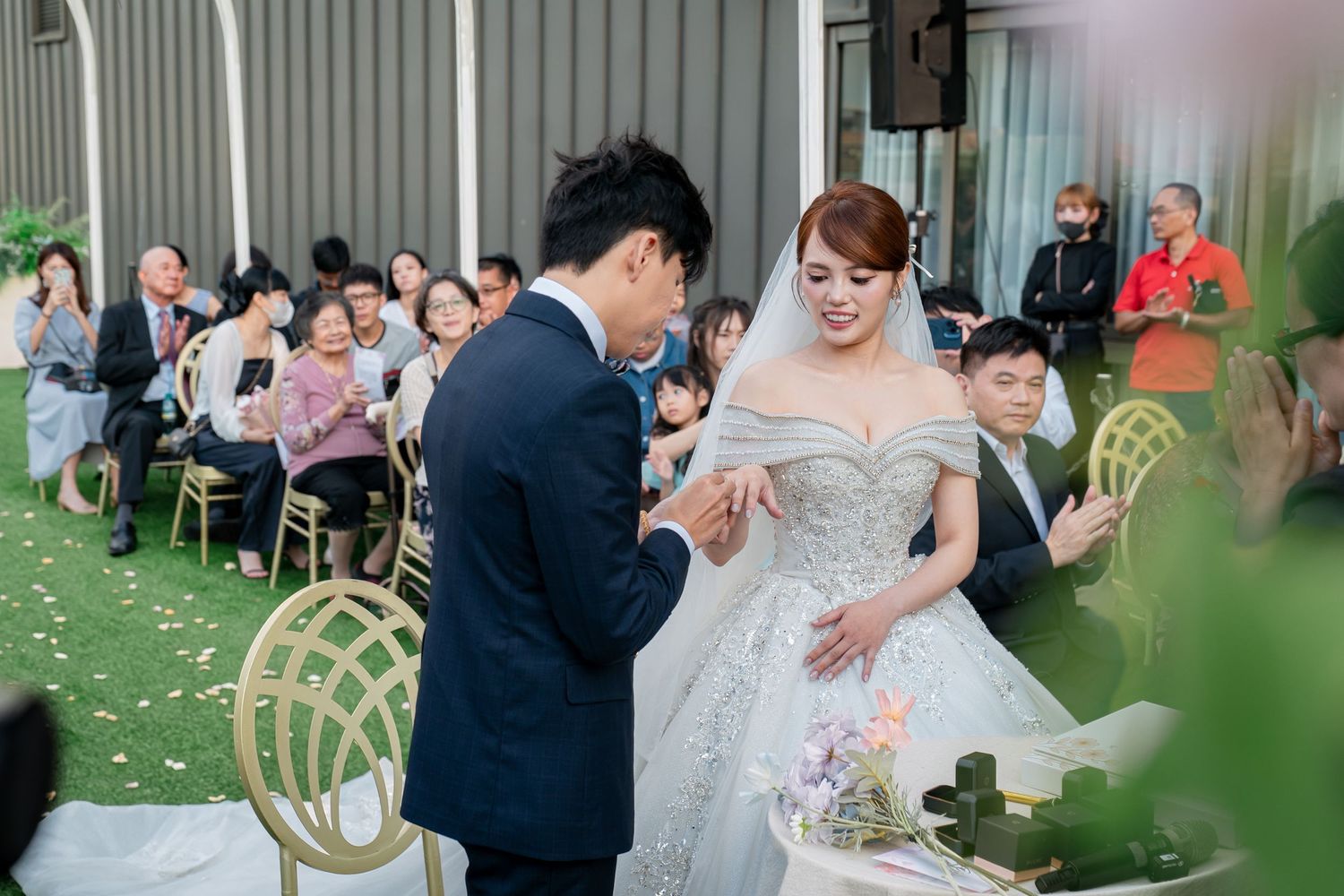 A groom places a ring on his bride's finger during an elegant wedding ceremony with guests seated in the background.