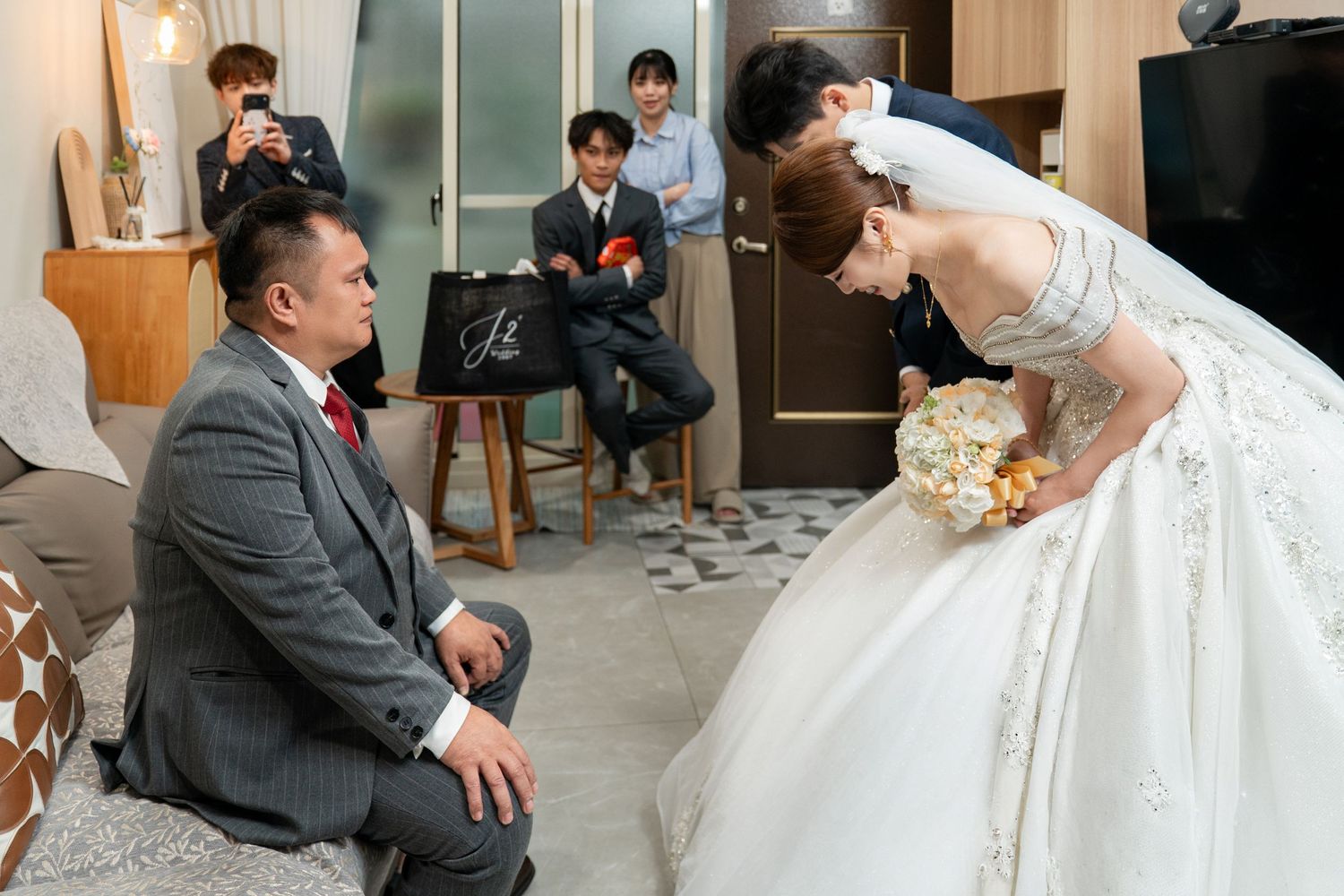 Bride in white gown bows to seated groom in gray suit during traditional wedding ceremony indoors.