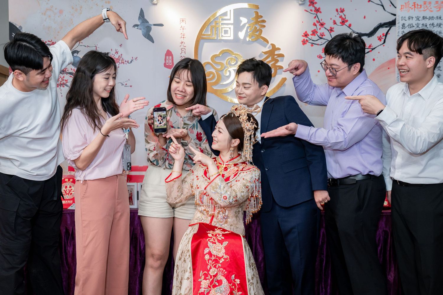 Group of friends in a festive room raising glasses and cheering around a bride in a red and gold traditional Chinese wedding dress.