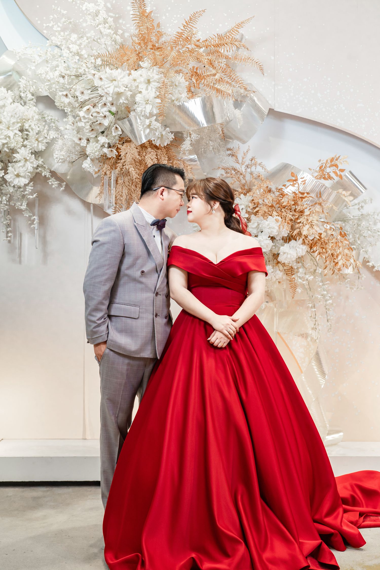 A couple in a grey suit and red ballgown embrace under white and gold floral decorations in an elegant indoor setting.