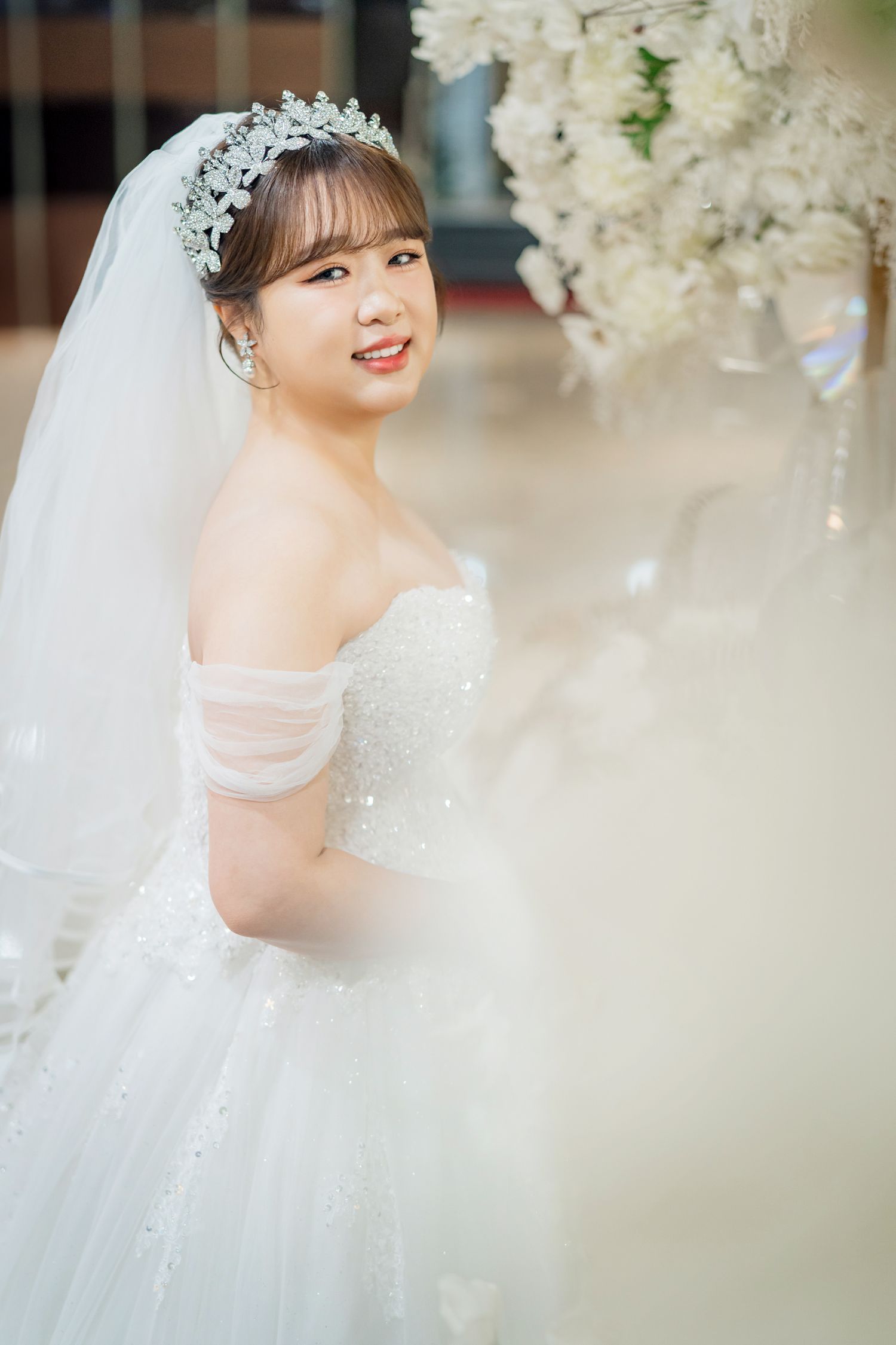 A bride in a white wedding dress and veil with a tiara stands elegantly among white floral decorations.