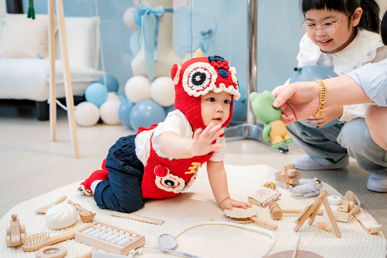 A baby in a red outfit plays with wooden toys on the floor while someone crouches nearby during a party celebration.