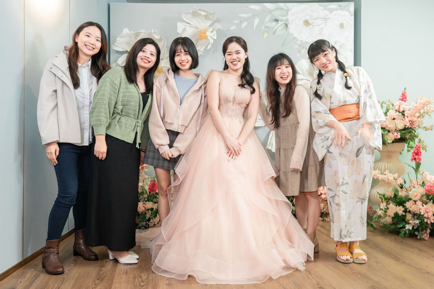 A group of friends pose together at a wedding celebration with one wearing a pink princess gown among floral decorations.
