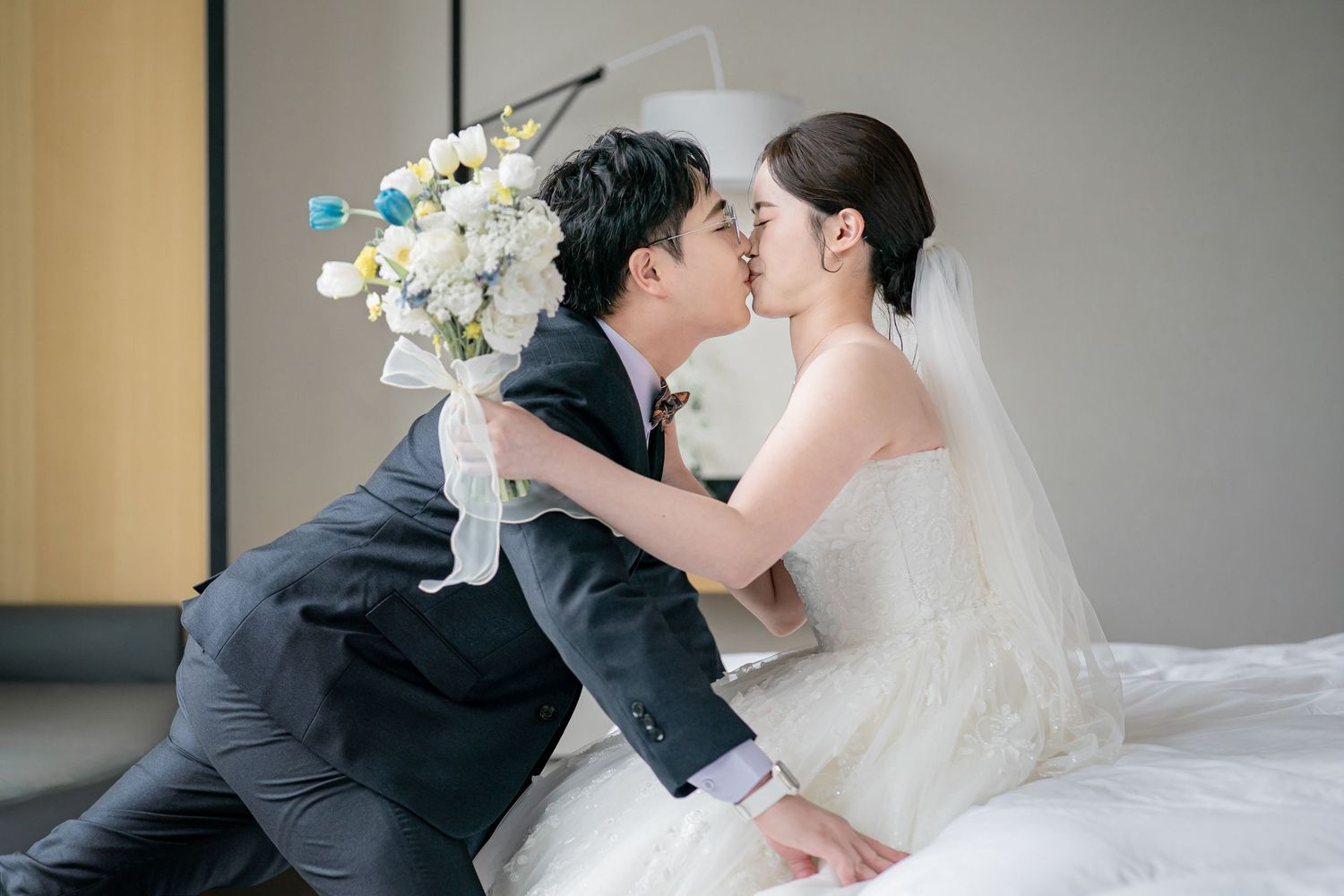 A wedding couple shares a romantic kiss while holding white floral bouquets in an elegant indoor setting.