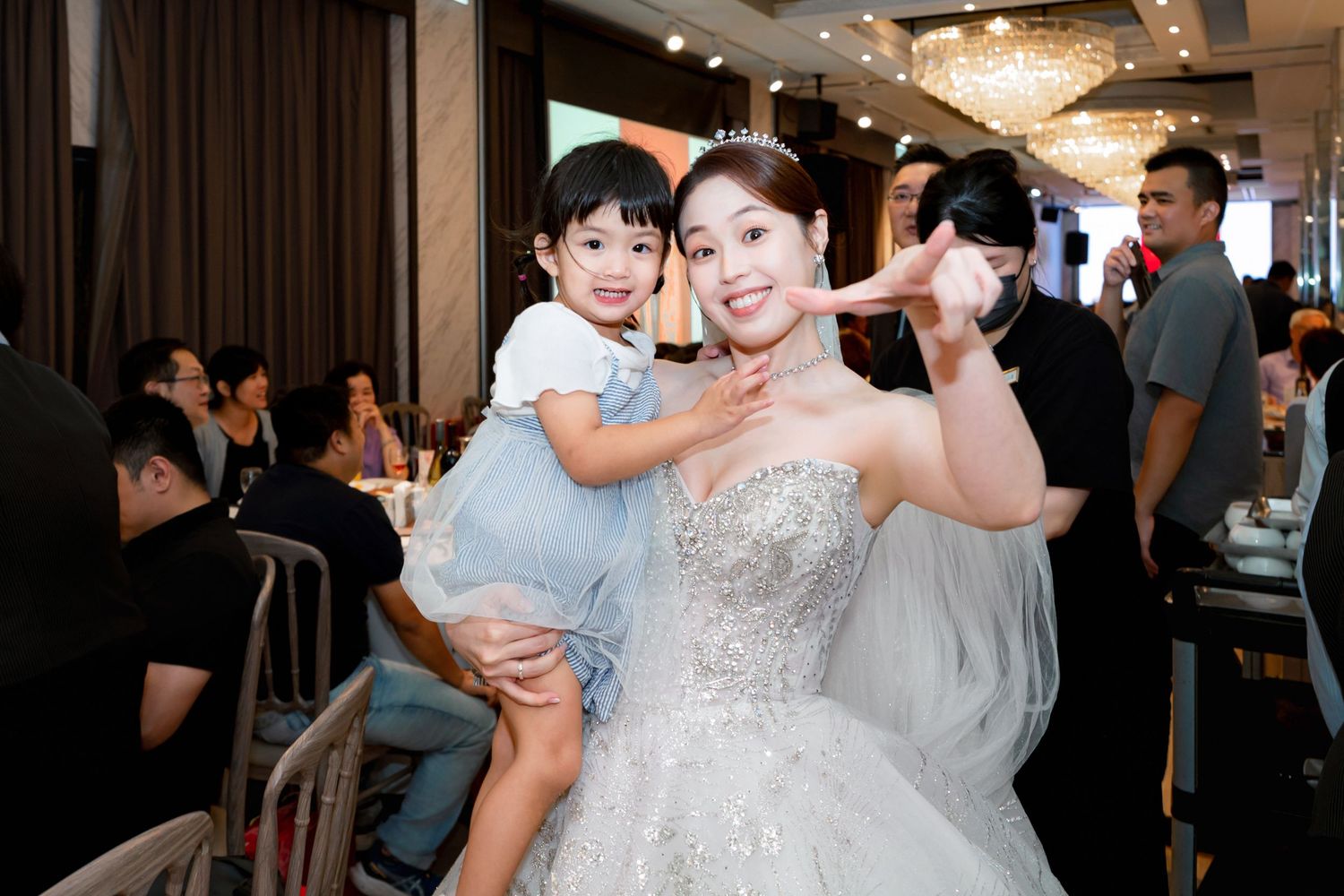 Bride in a sparkly white gown dances with guests at a wedding reception under elegant chandelier lighting.