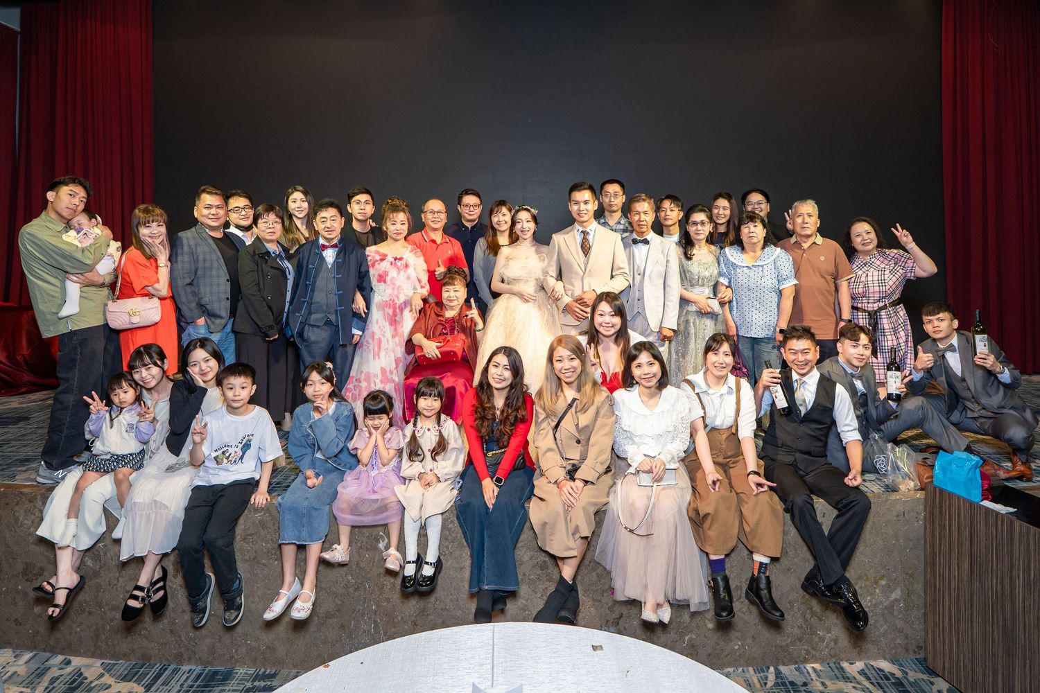 Large group of formally dressed Asian adults and children posing together at a wedding celebration indoors.