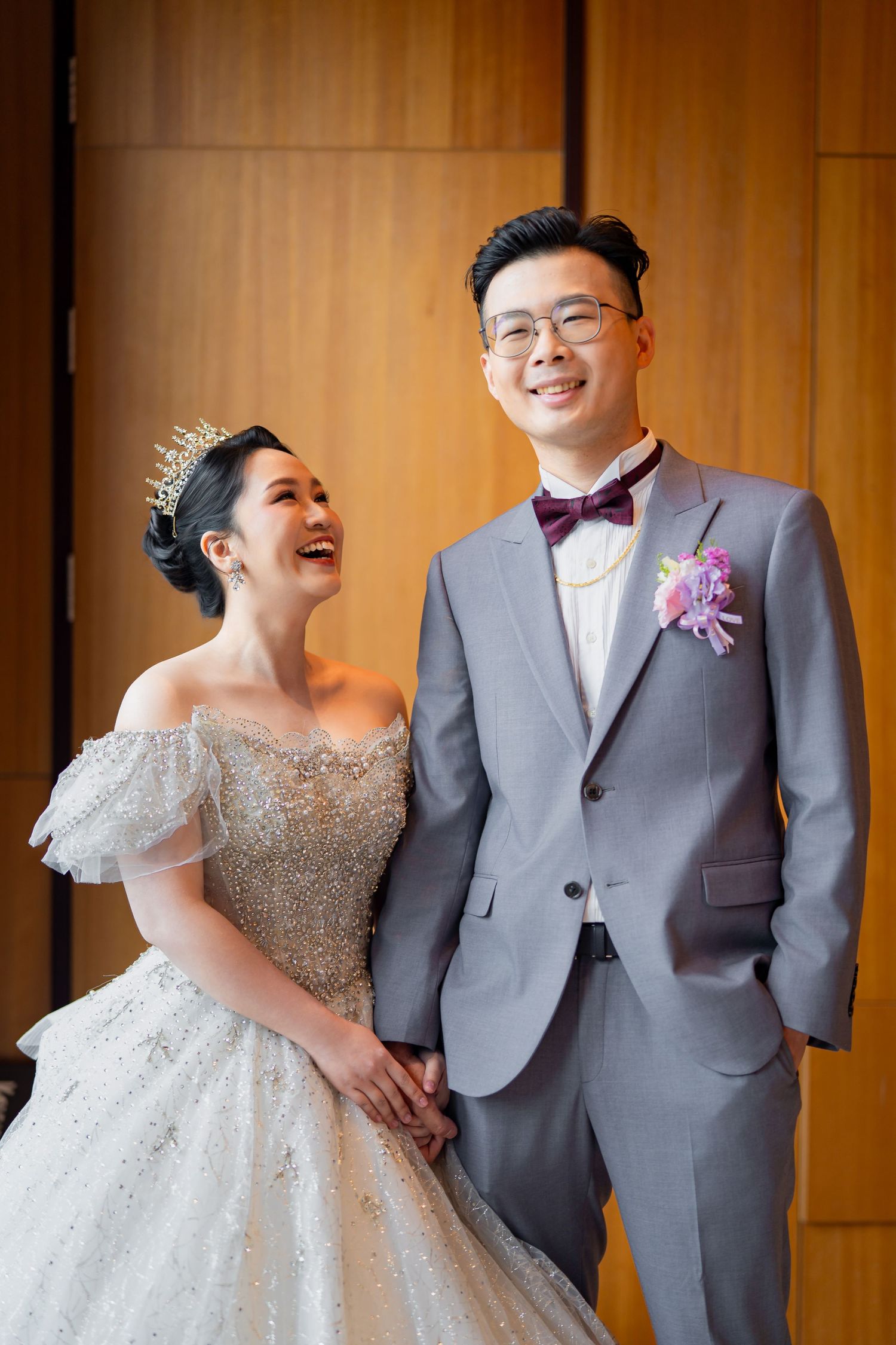 A happy couple in wedding attire share a candid moment against a wooden backdrop, with bride in white gown and groom in grey suit.