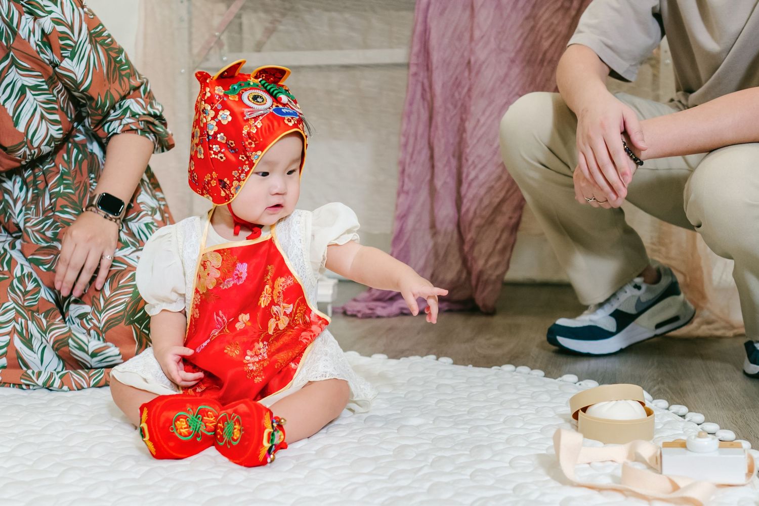Small child wearing vibrant red traditional Asian dress plays with blocks on a white carpet during playtime.