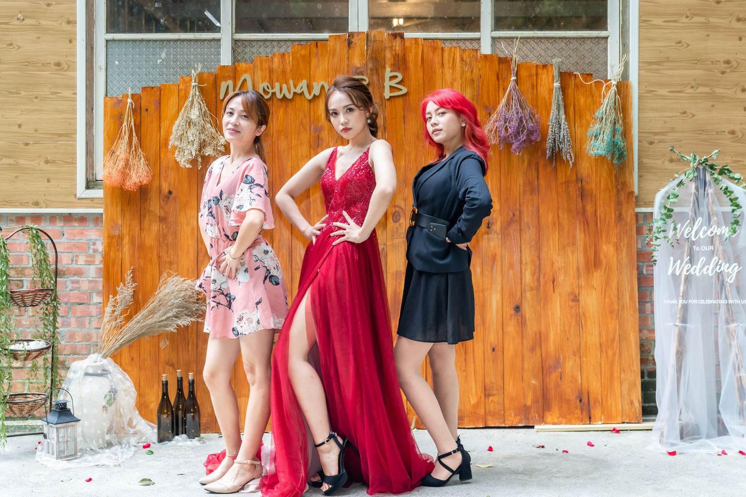 Three models in contrasting dresses pose together against a rustic wooden wall with hanging decorations.