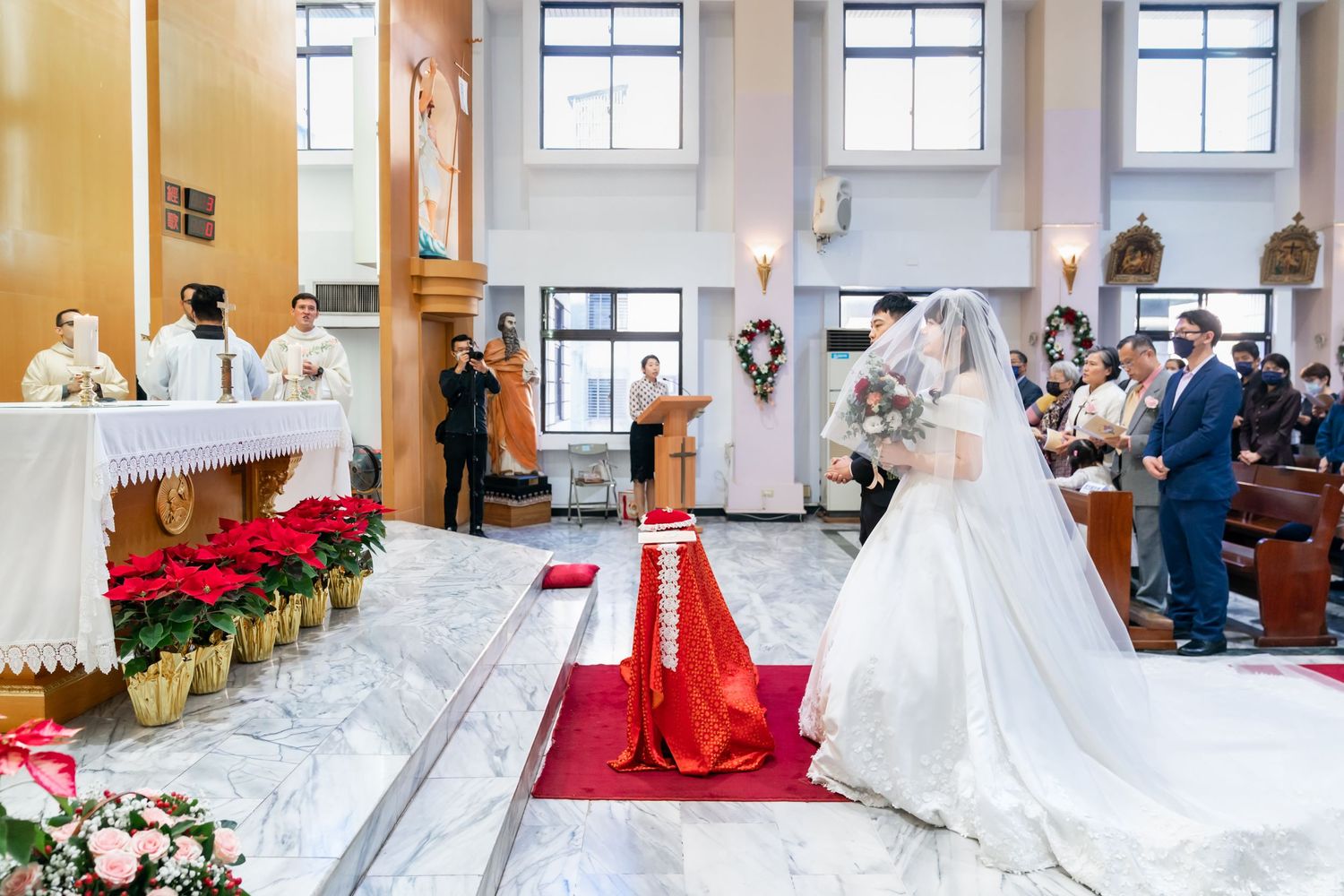 Church wedding ceremony with bride in flowing white gown and red carpet leading to decorated altar.