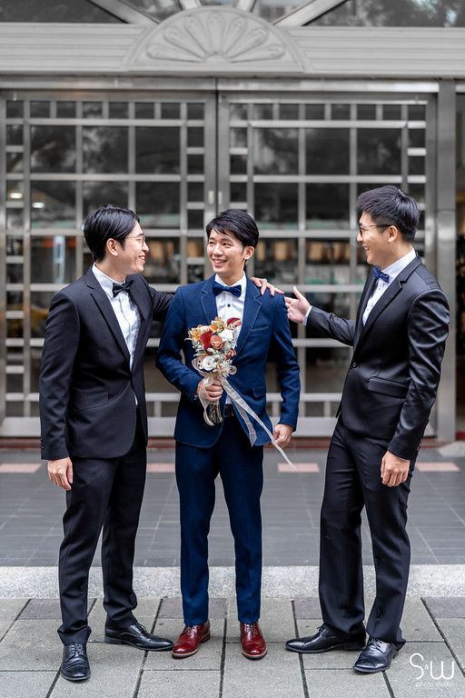 Groomsmen in black suits share a laugh with groom in navy suit holding bouquet outdoors.