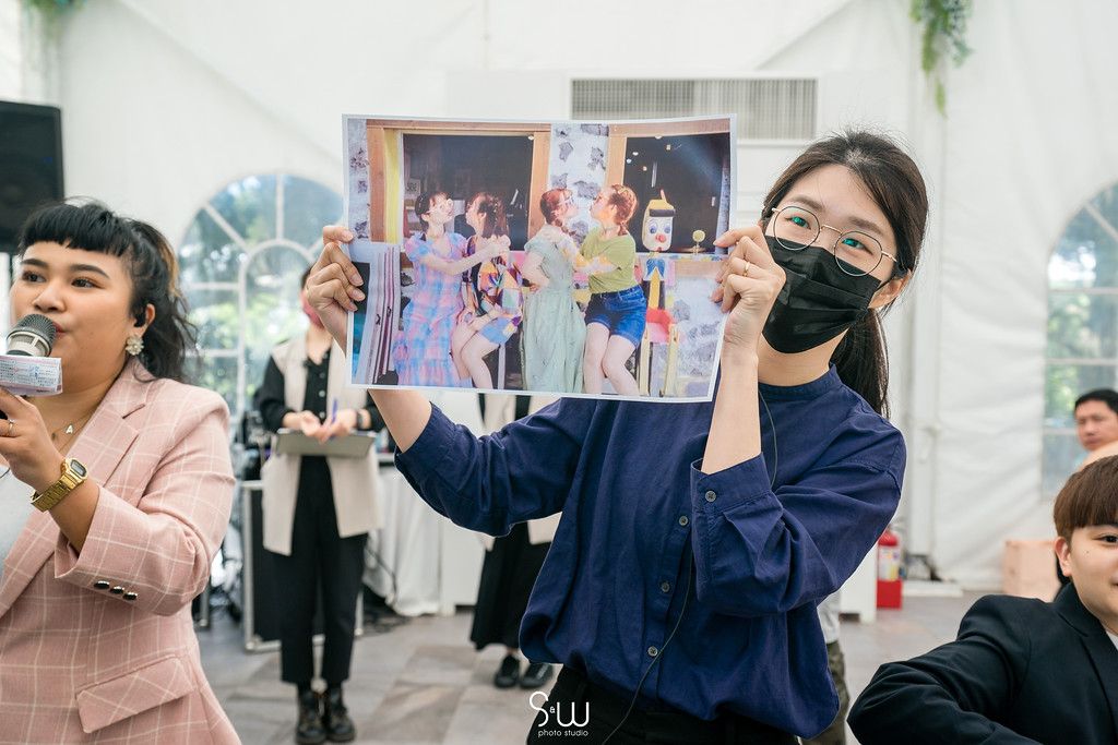 Wedding guests in masks hold up photographs during a celebratory gathering at an indoor venue.