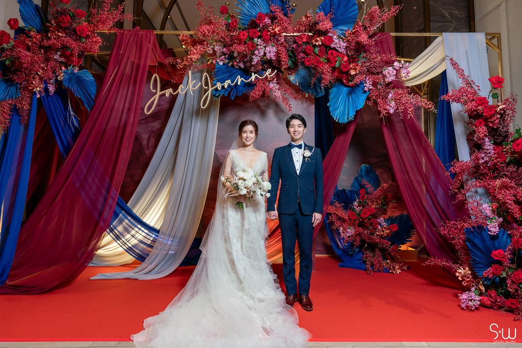 Elegant wedding backdrop features red cherry blossoms and blue draping with a couple on red carpet.