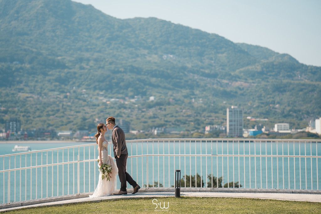 Wedding couple stands on waterfront balcony with scenic mountain and ocean view in background.