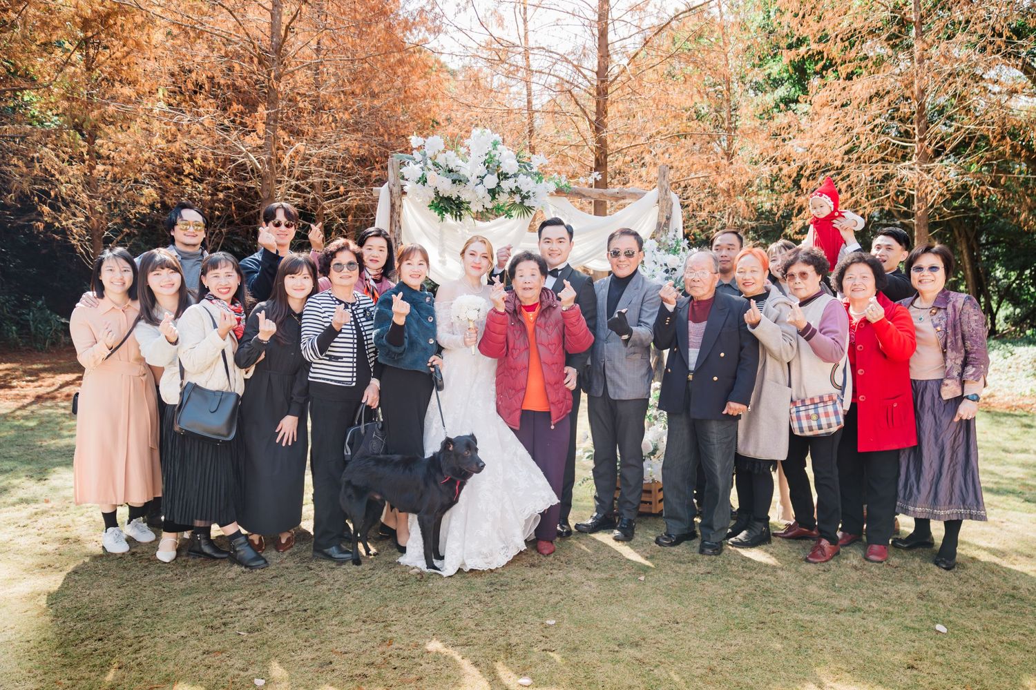 Large group photo of wedding party and guests beneath autumn trees.