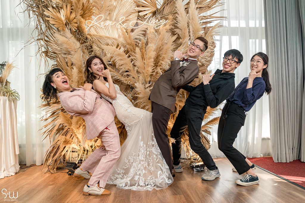 Fun and playful wedding party pose with dramatic expressions against pampas grass wall.