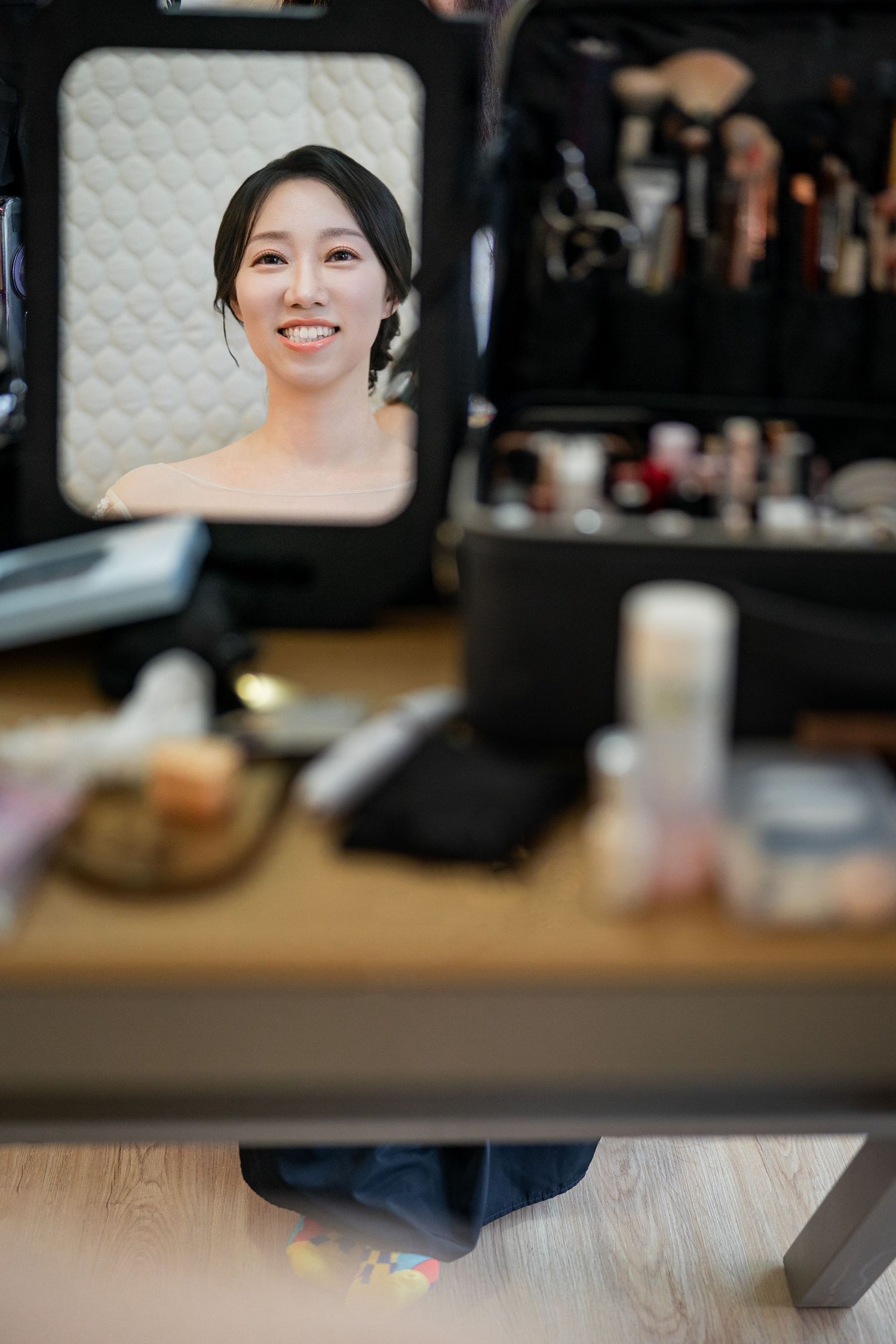 A reflection in a makeup mirror shows a smiling expression with makeup products visible on the wooden table.