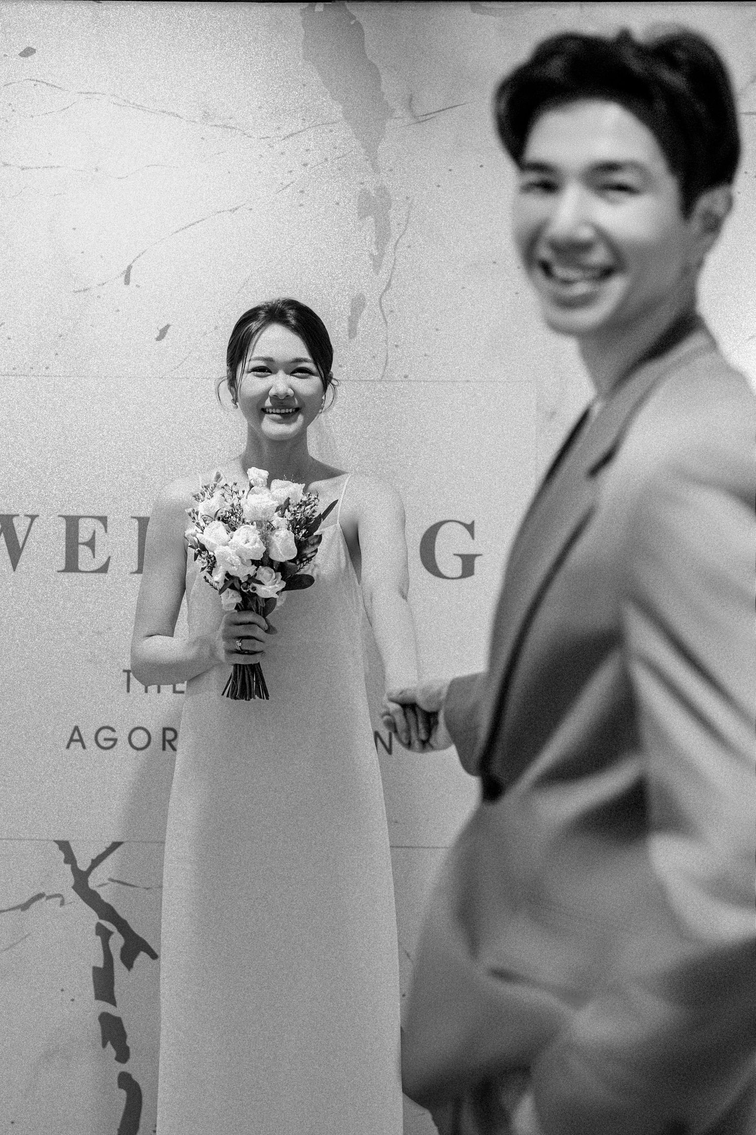 A black and white wedding photo showing a bride in a long gown holding a bouquet while smiling next to a suited groom.