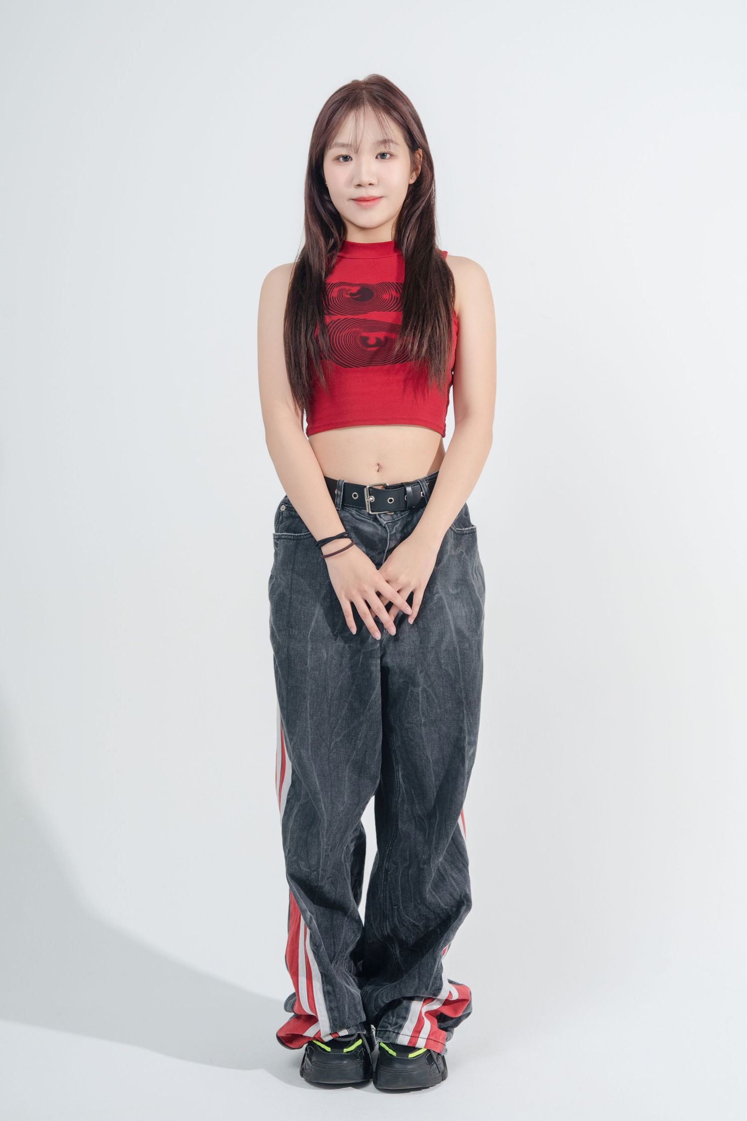 Person wearing red crop top and baggy jeans crouches against white background in casual urban style pose.
