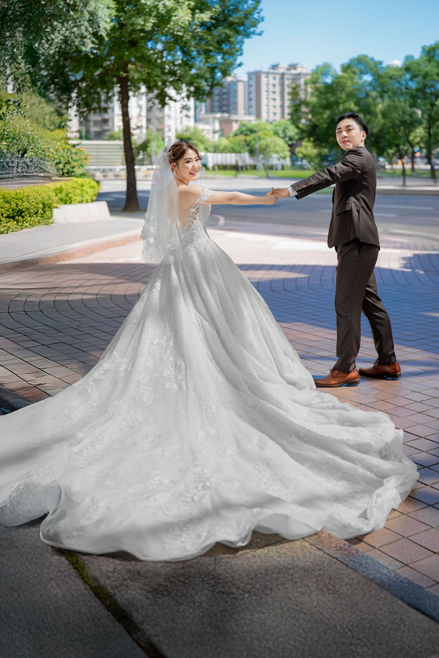 A wedding couple poses outdoors on a brick paved walkway with a flowing white lace gown spread across the ground.
