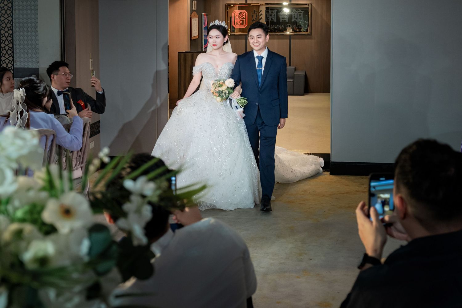 A wedding couple walks down a hallway while photographers capture their special moment.
