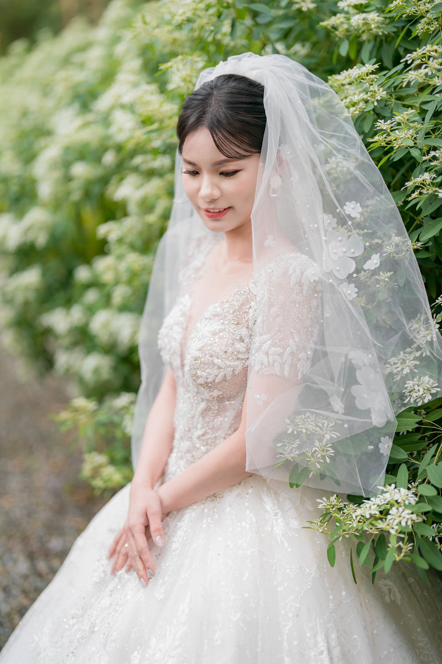 A bride in an elegant white lace wedding gown and veil poses against a backdrop of lush green foliage.