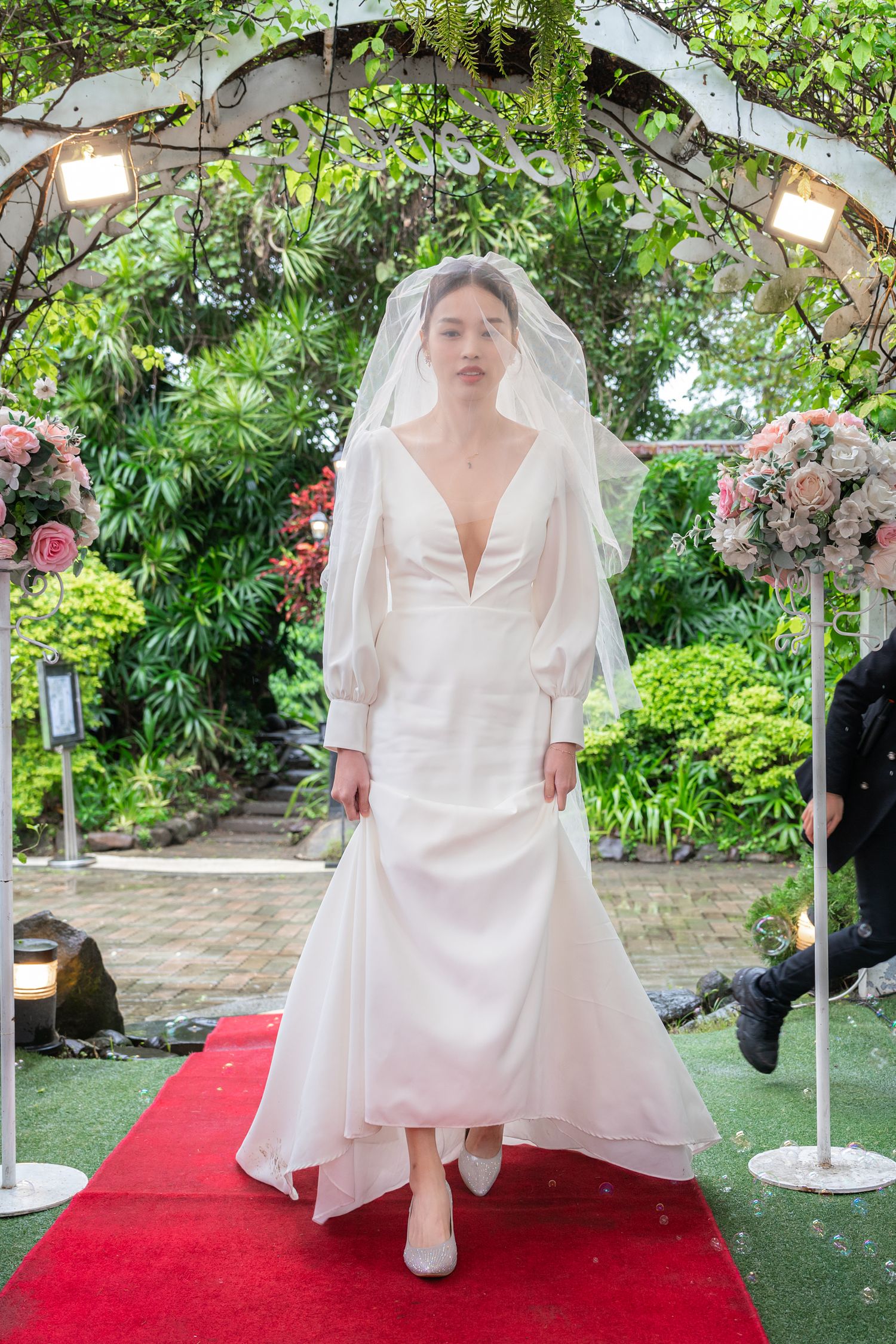 Elegant white wedding dress with long sleeves and plunging neckline on red carpet under floral garden arch with lights.