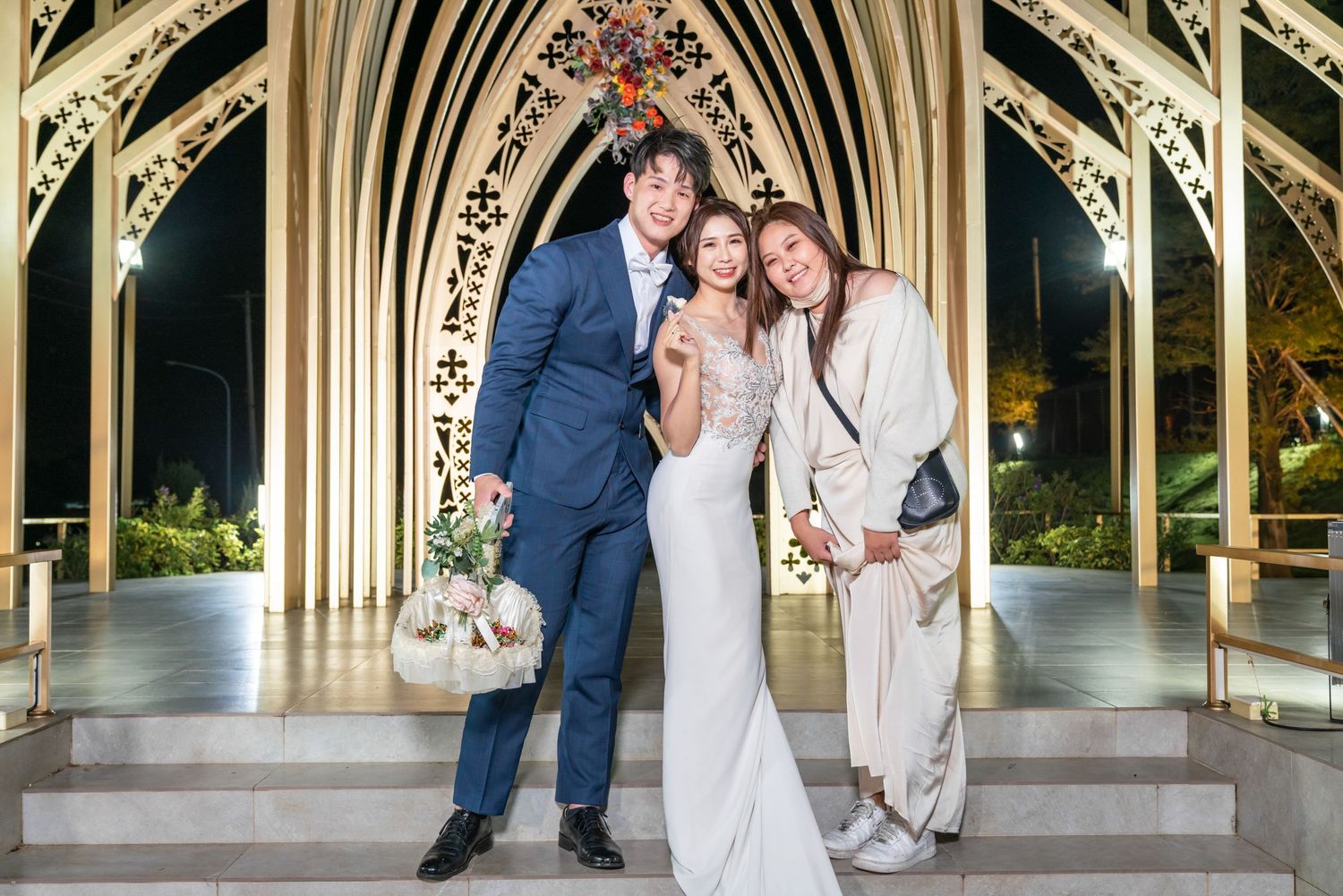 Three people pose together in formal attire under an ornate Gothic archway at a nighttime wedding celebration.