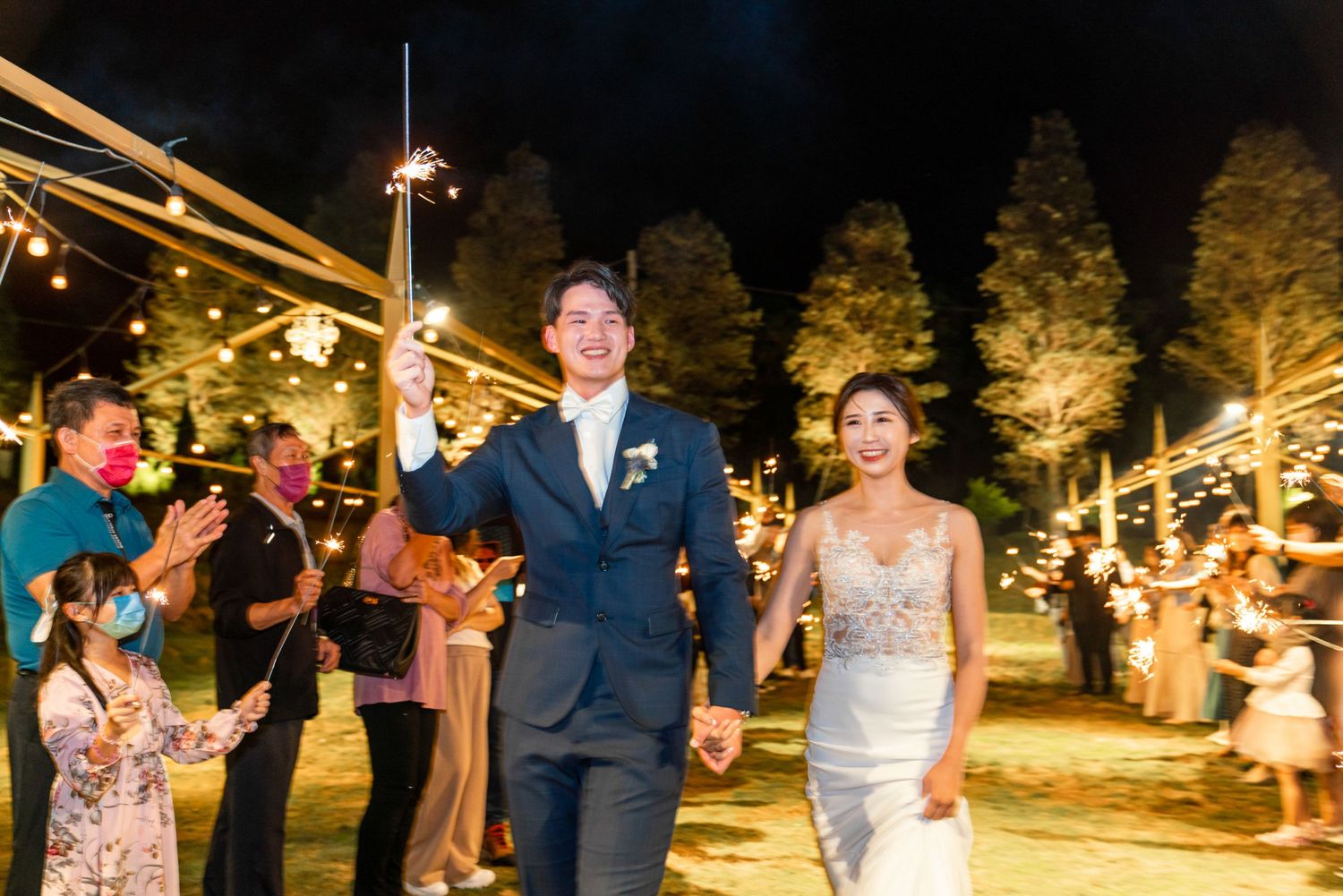 A happy couple celebrates at an outdoor evening wedding reception beneath glowing string lights.