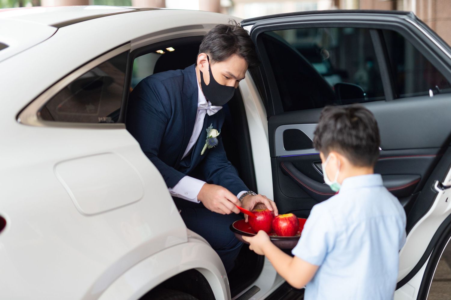 A person in a black suit and face mask passes keys to another person by a car door.