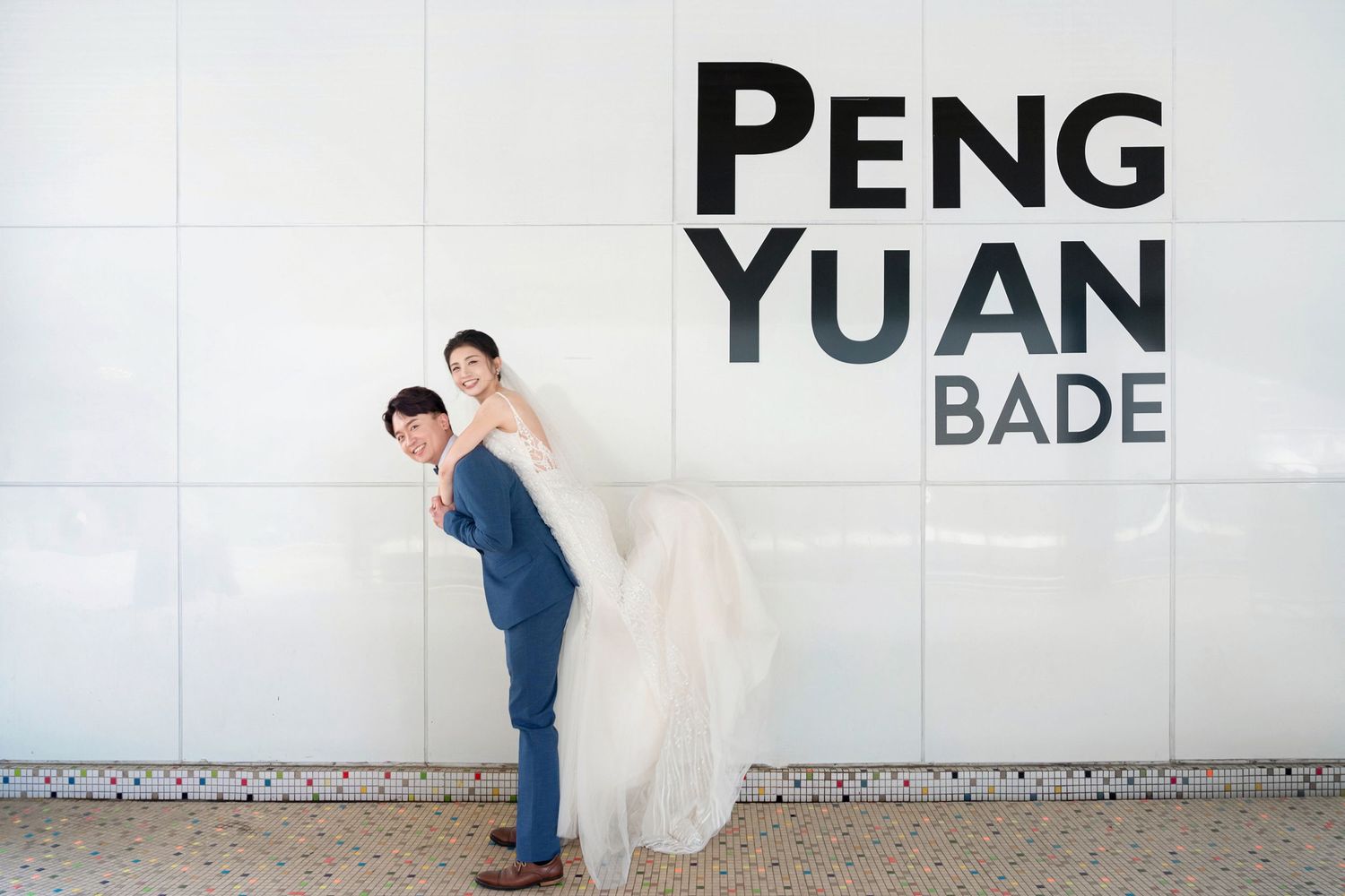 A couple poses in front of a wall with text reading 'PENG YUAN BADE', with the bride's dress flowing elegantly.
