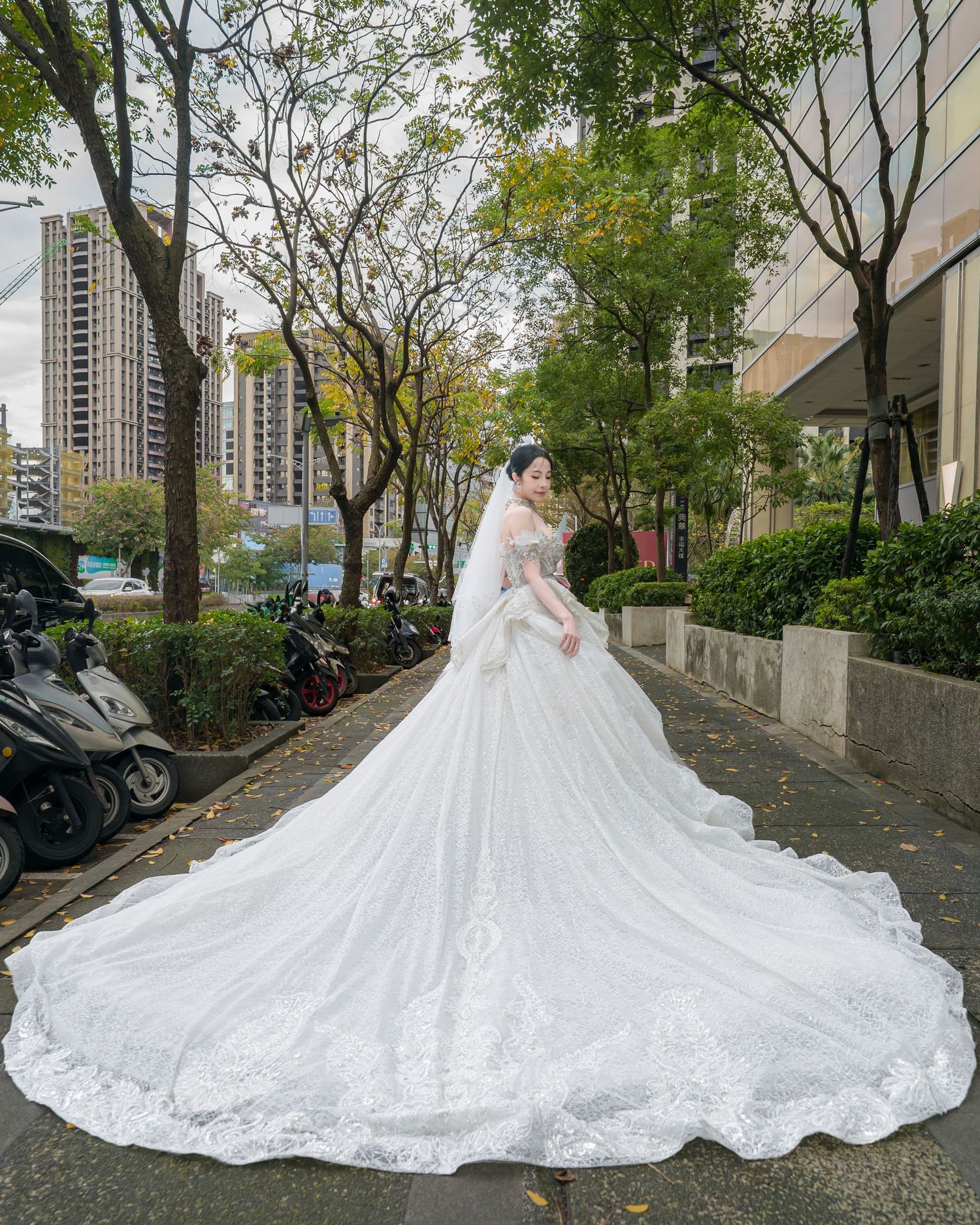 A stunning bridal gown with an elaborate cathedral-length train flows across a tree-lined urban pathway.
