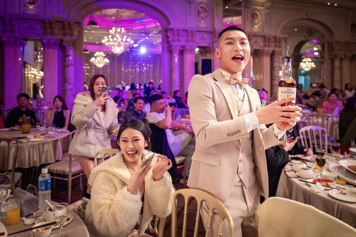 A group of wedding guests dancing and celebrating in a luxuriously decorated ballroom with purple lighting.