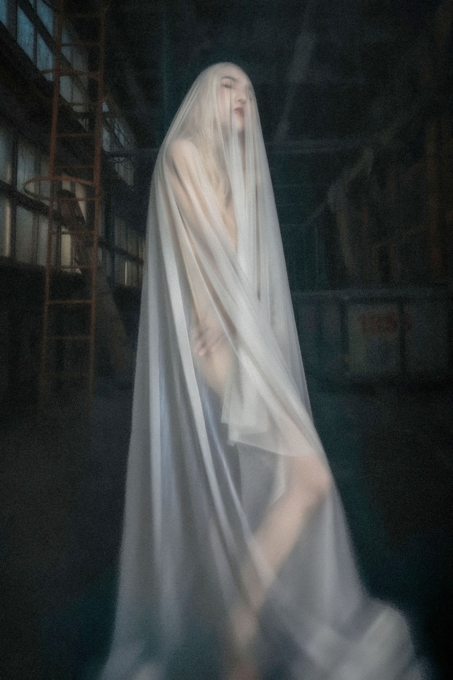 Ethereal figure draped in flowing white fabric stands mysteriously in a dark, moody indoor setting creating a ghostly atmosphere.