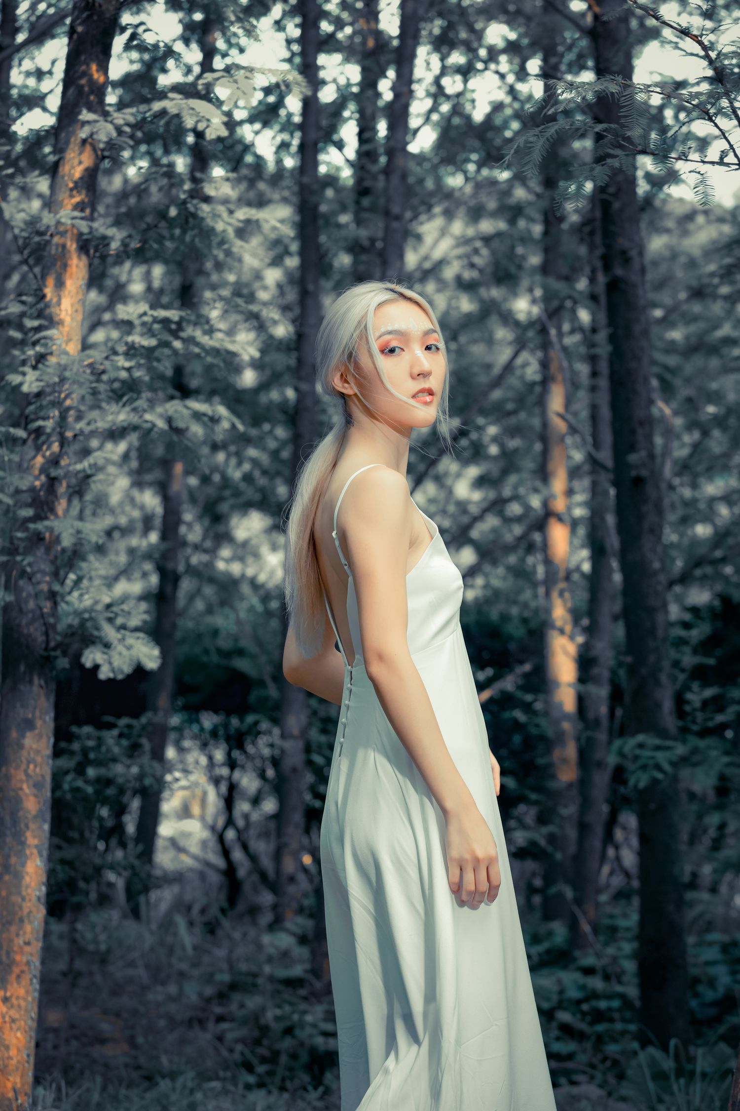 A figure in an ethereal white dress poses elegantly in a misty forest setting with sunlight filtering through the trees.