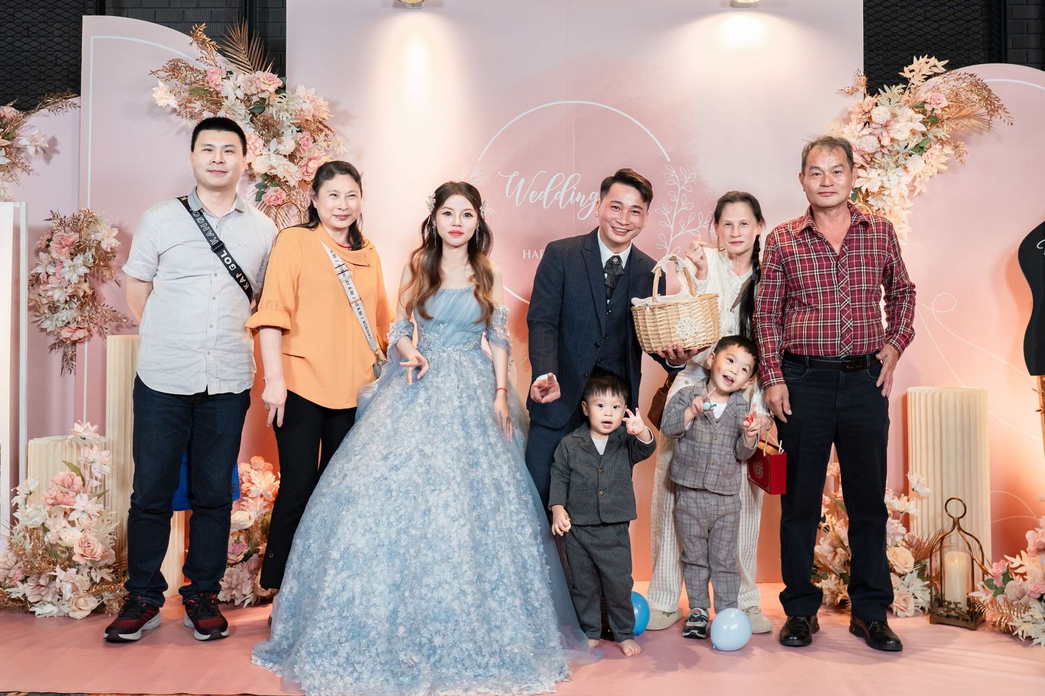 A group of wedding guests gather around a couple in formal attire at an elegant pink and floral decorated venue.