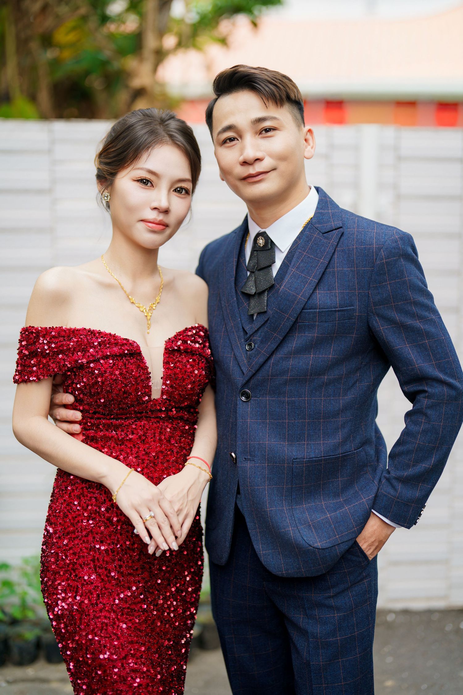 A couple poses in formal attire, with a red sequined gown and navy blue suit against a light backdrop.