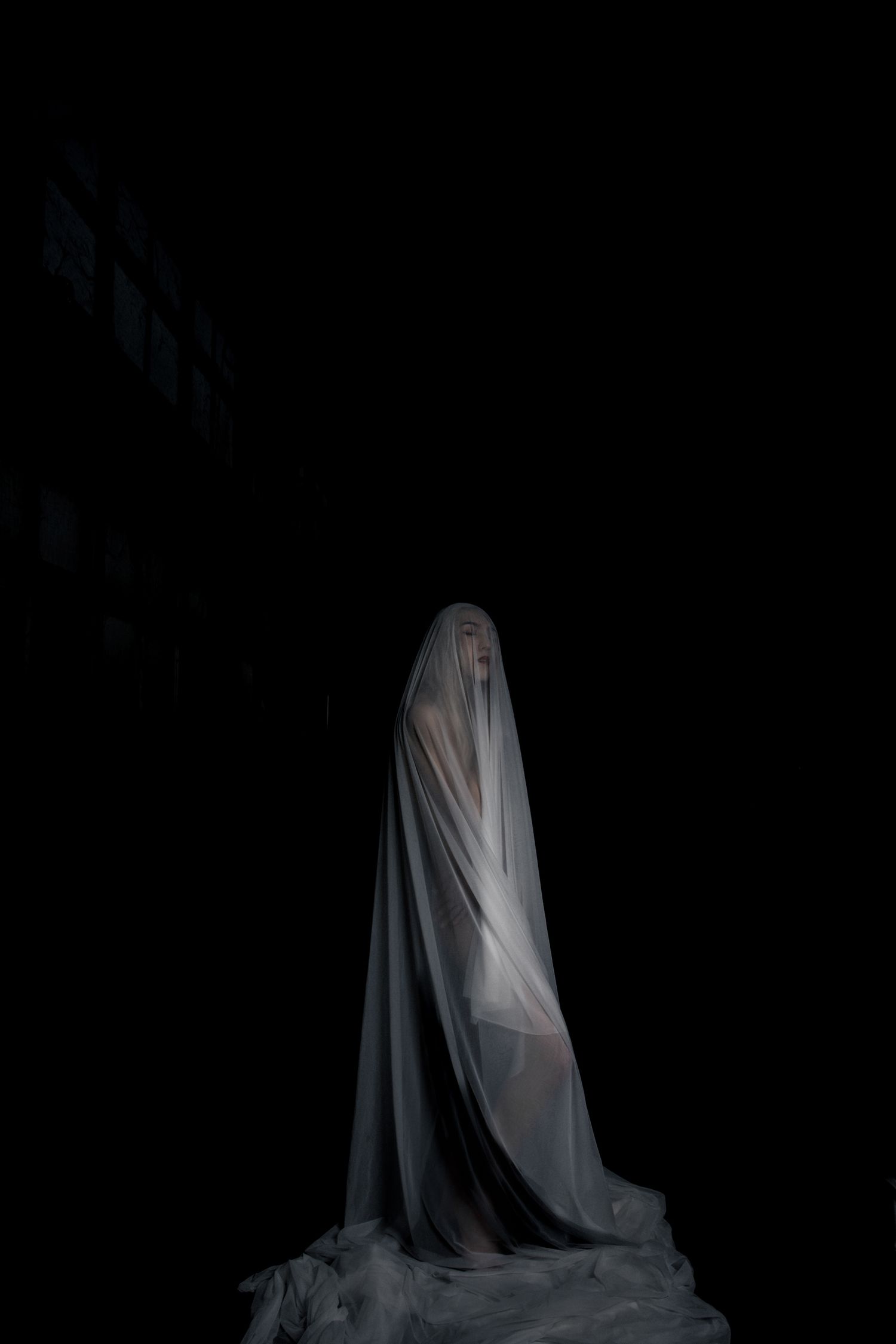 A ghostly figure draped in white translucent fabric stands against a dark black background in eerie fine art photography.