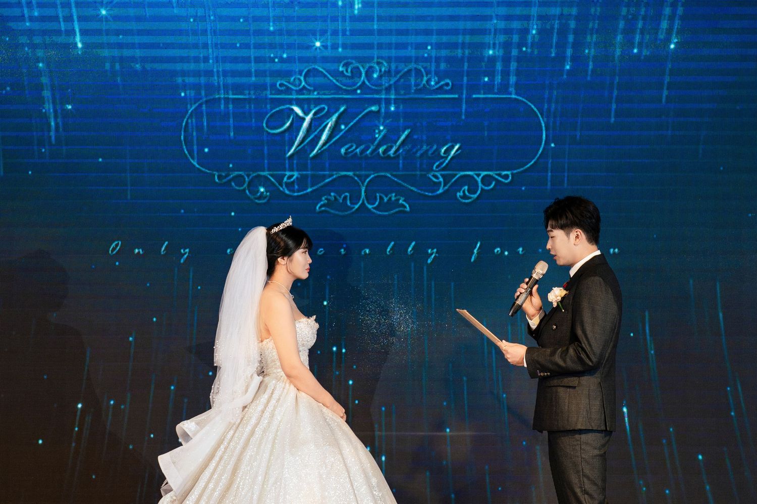 Bride in white gown and groom in black suit exchange vows on a blue-lit wedding stage.