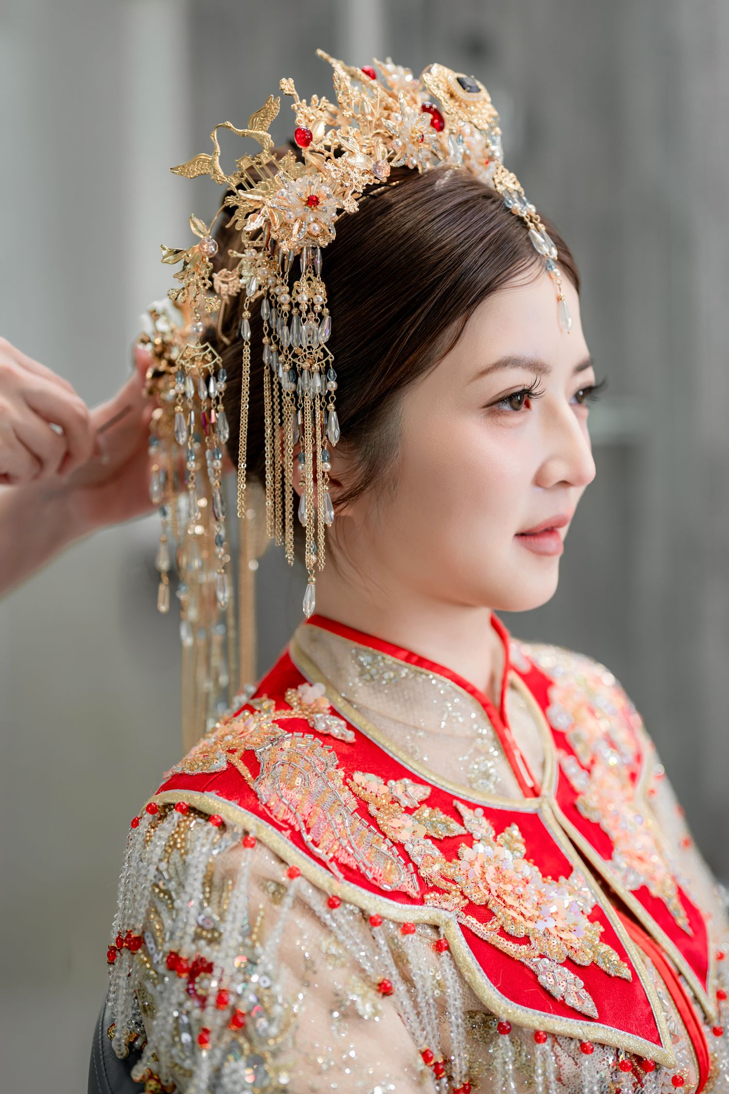 Bride in a red and gold embroidered Chinese wedding gown and ornate beaded phoenix headdress as hands adjust her hair before the ceremony.