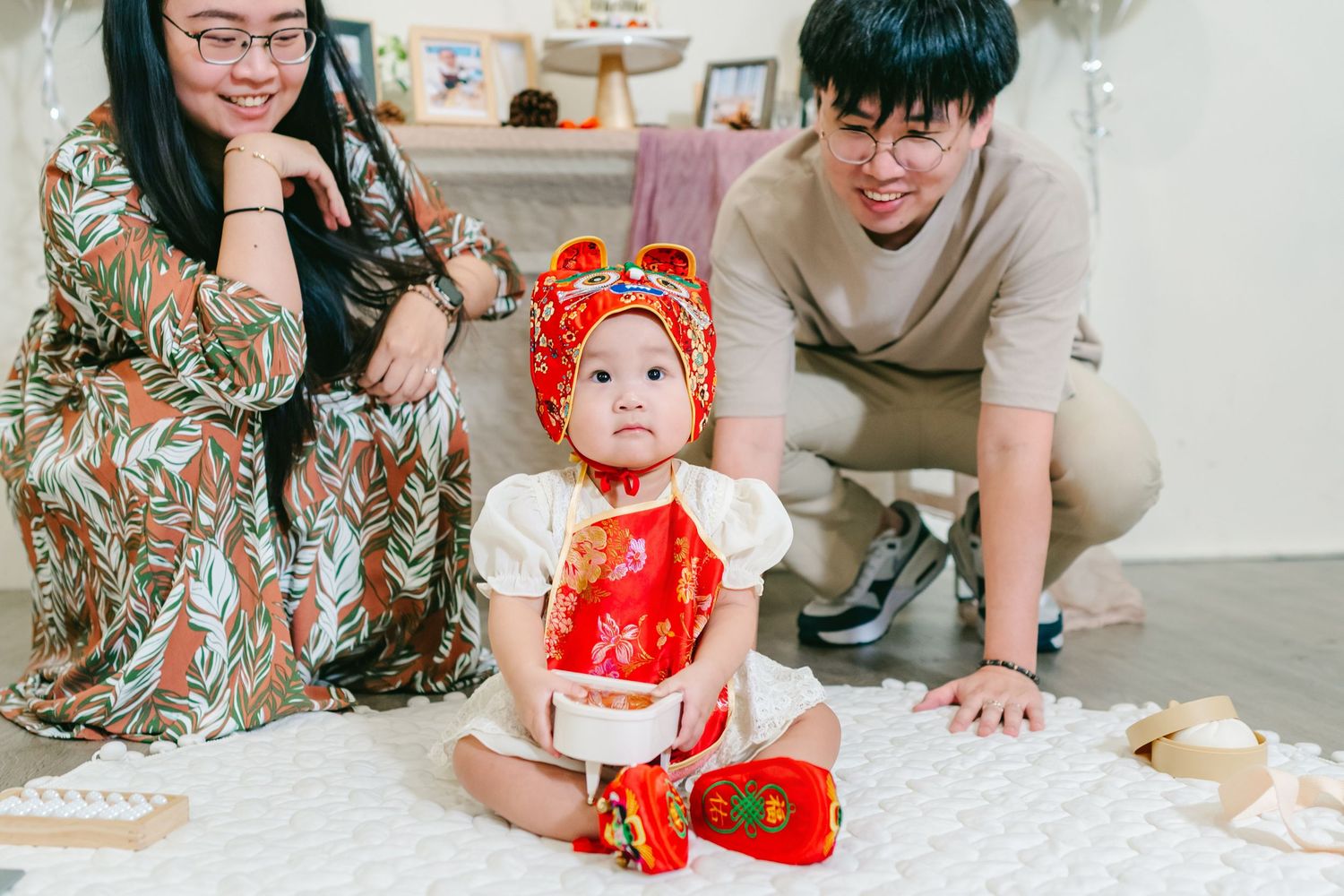 A family gathers around a traditional Chinese red and gold dress decorated cake during a first birthday celebration.