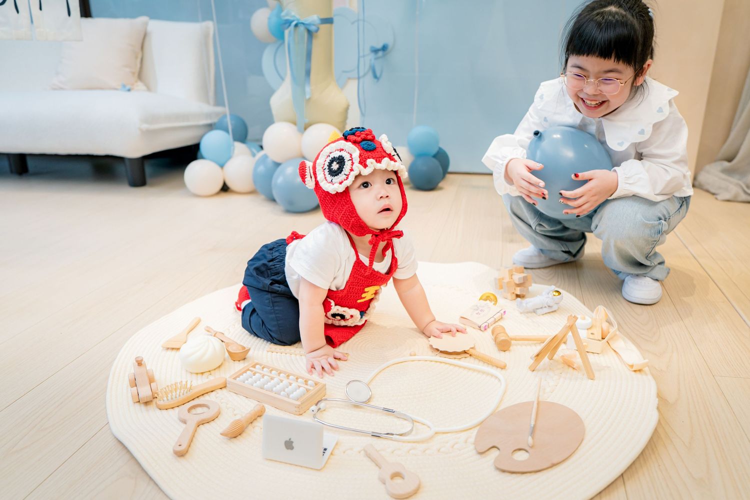 A baby in a red outfit crawls and plays on a white mat with various toys and stuffed animals spread around.