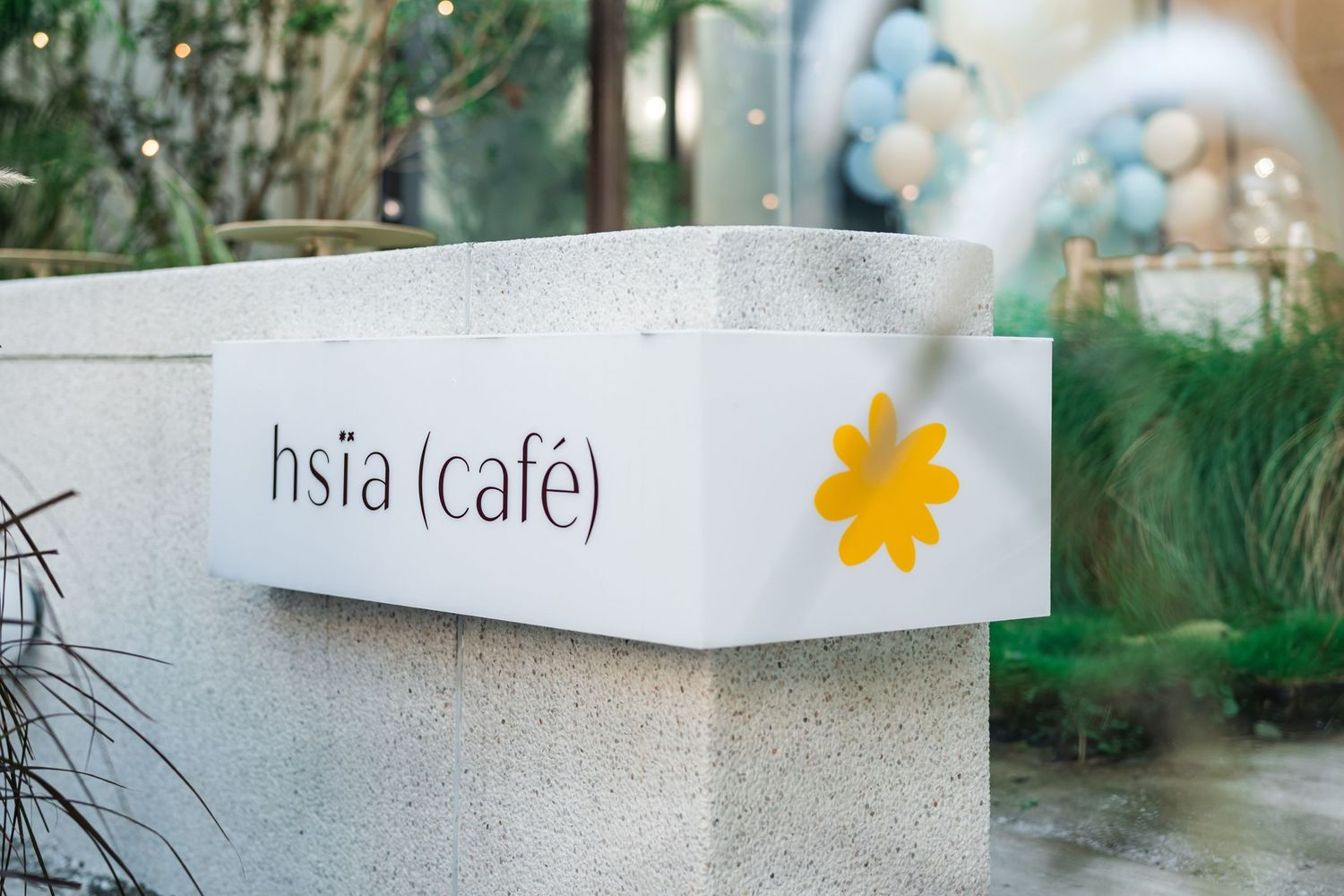 A minimalist white cafe sign with 'hsia (cafe)' text and a yellow flower design mounted on a concrete wall.