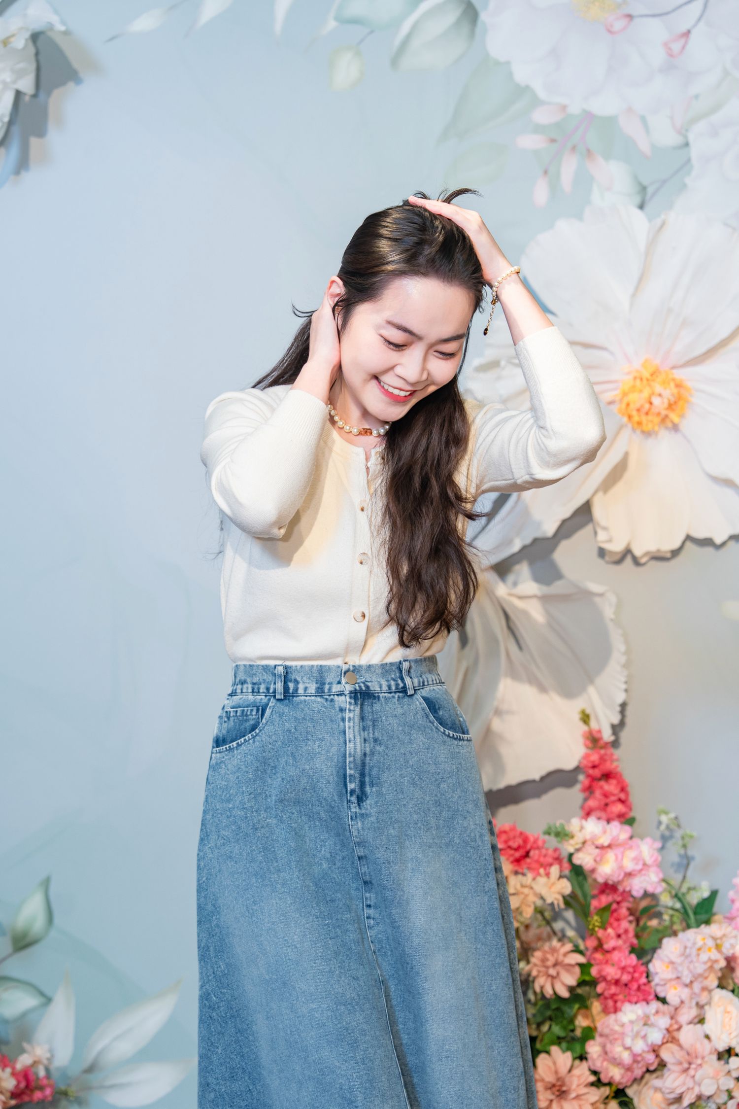 A woman in a cream sweater and jeans poses playfully against a floral backdrop in a spring-themed photoshoot.