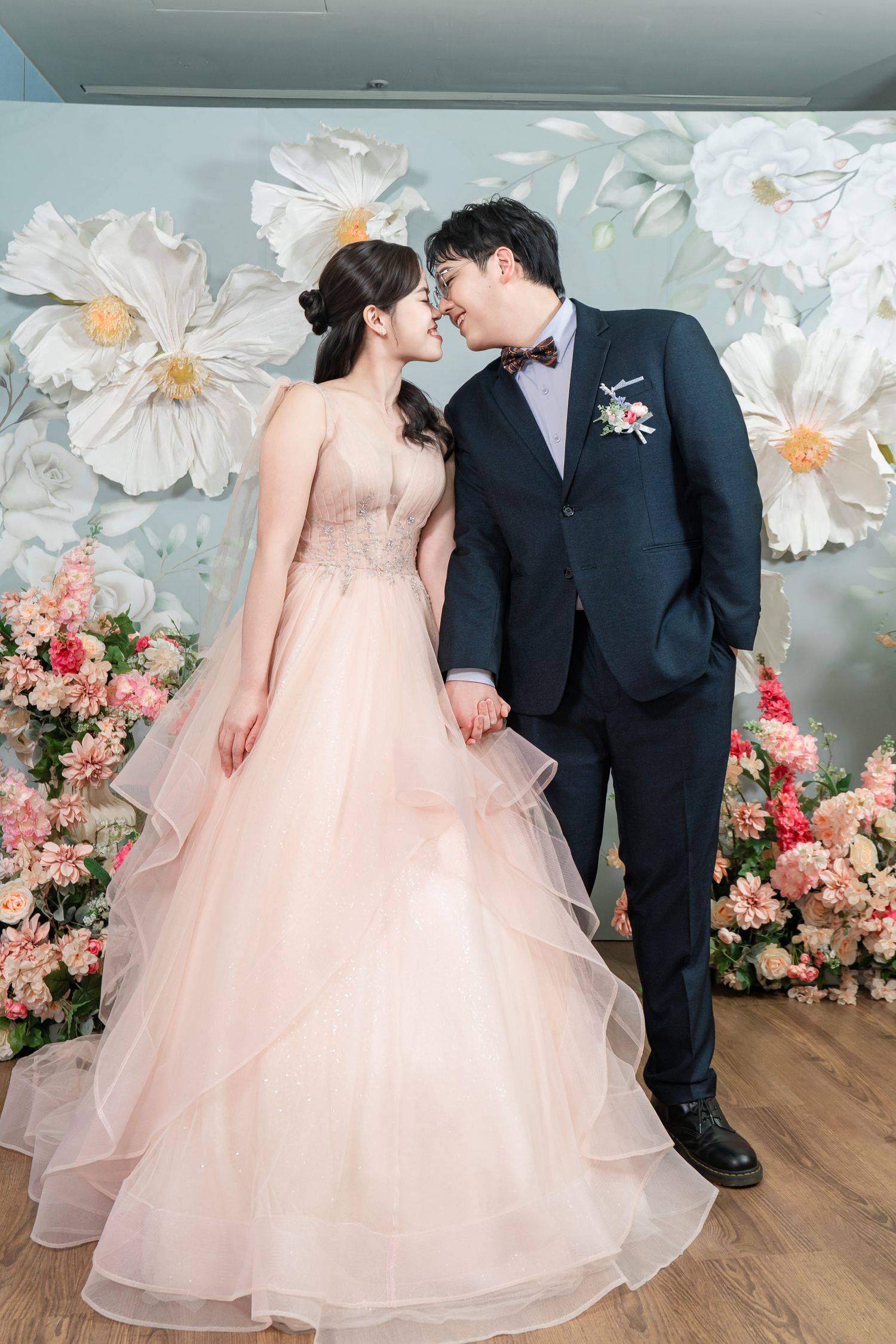 Couple in wedding attire share romantic moments against a floral backdrop with white paper flowers and pink roses.