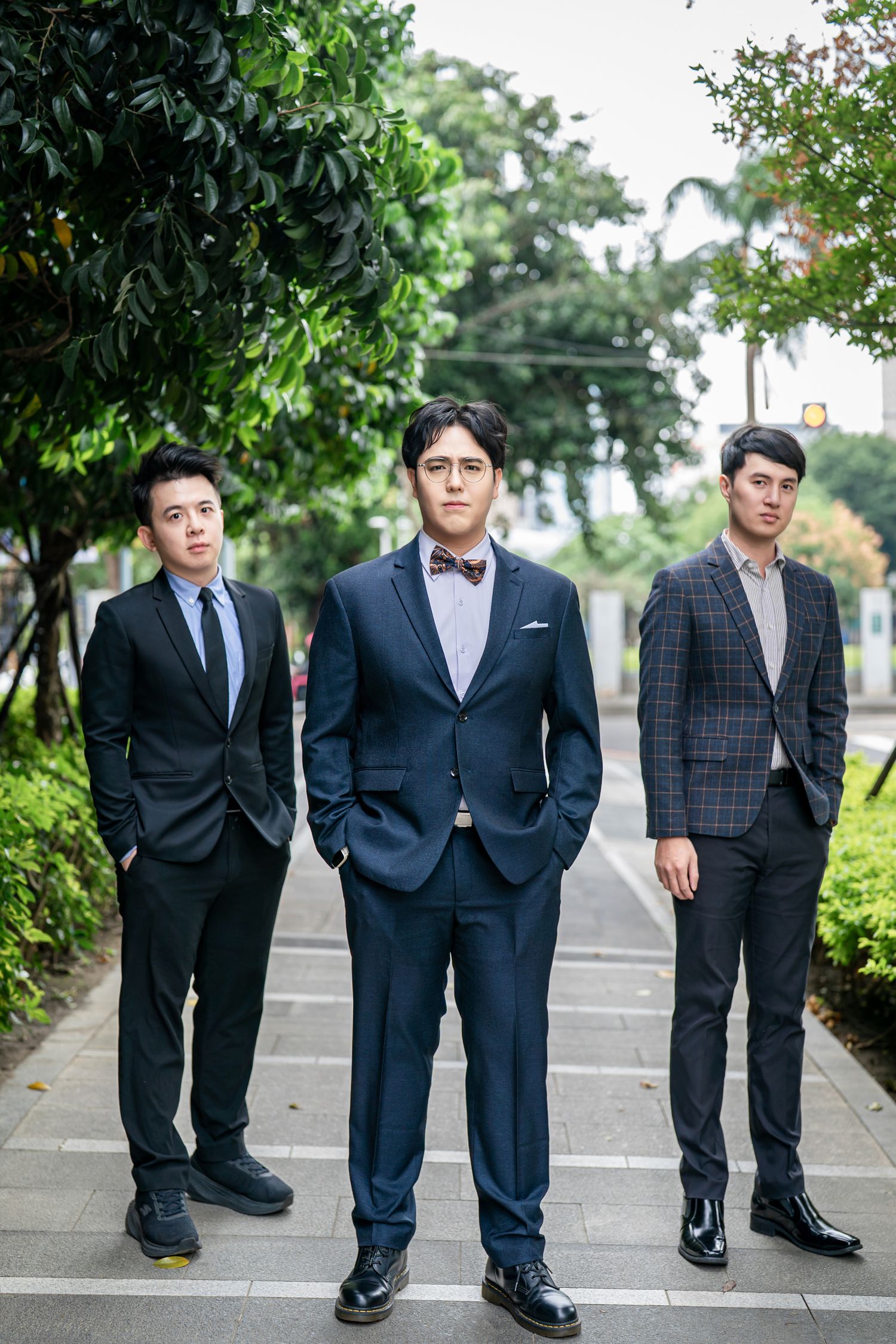 Three men in formal business suits and dress shoes stand on a tree-lined walkway outdoors.
