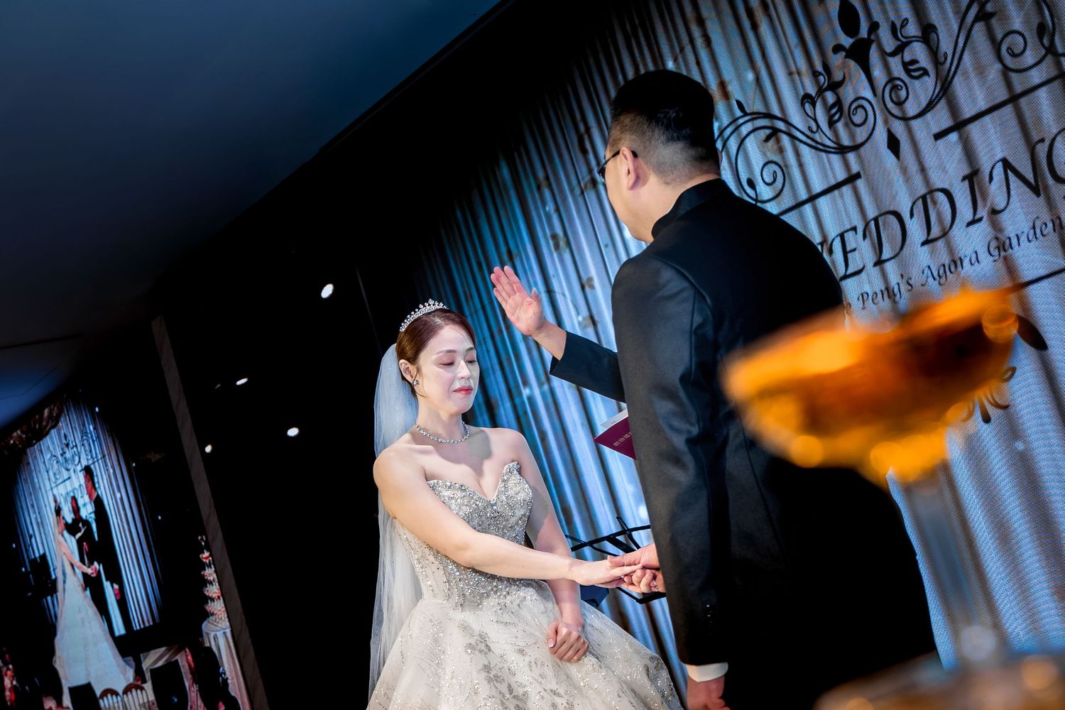 A couple in elegant wedding attire poses in front of a dark backdrop with dramatic lighting and text elements.