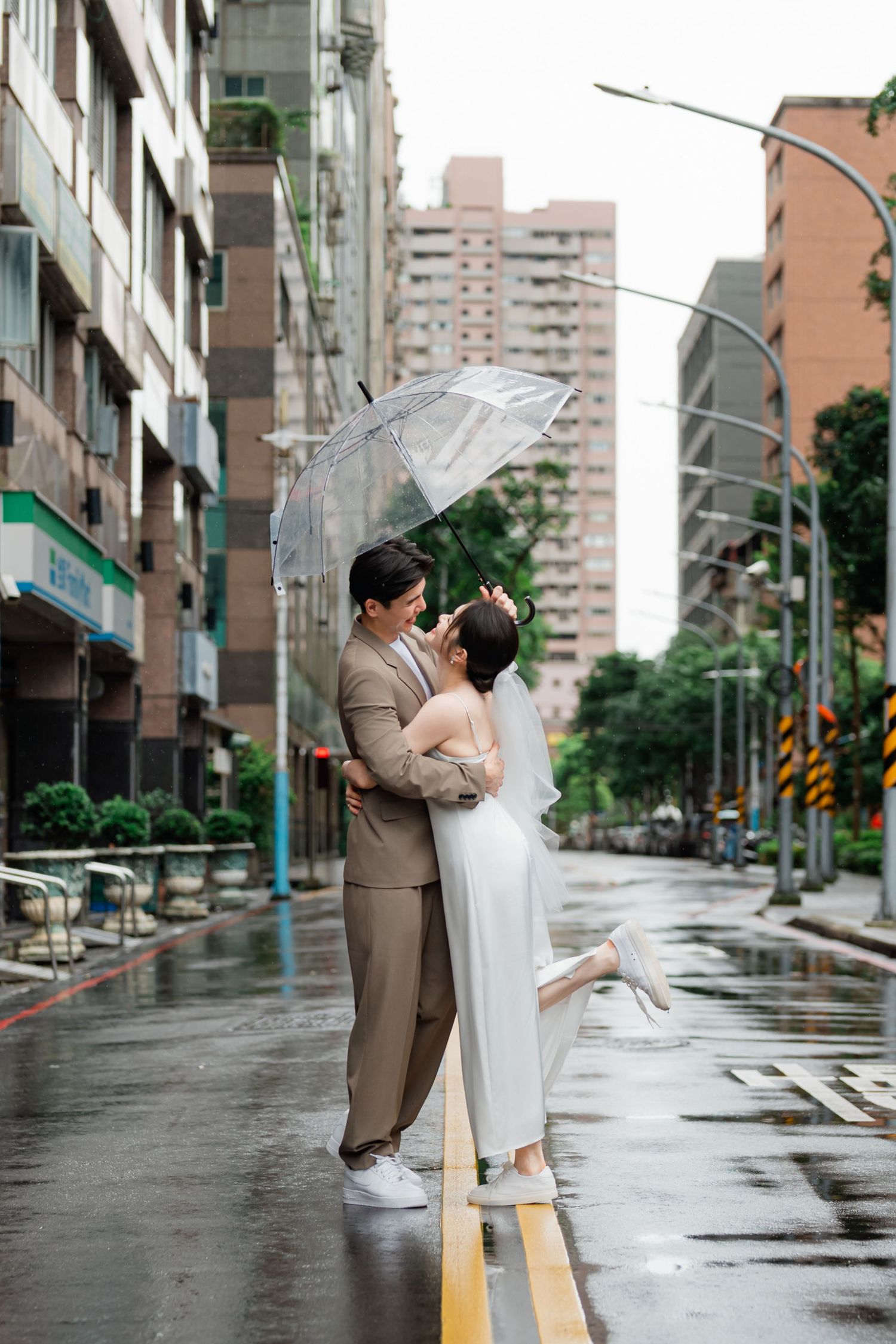 A romantic moment shared under a clear umbrella on a rainy city street with high-rise buildings in the background.