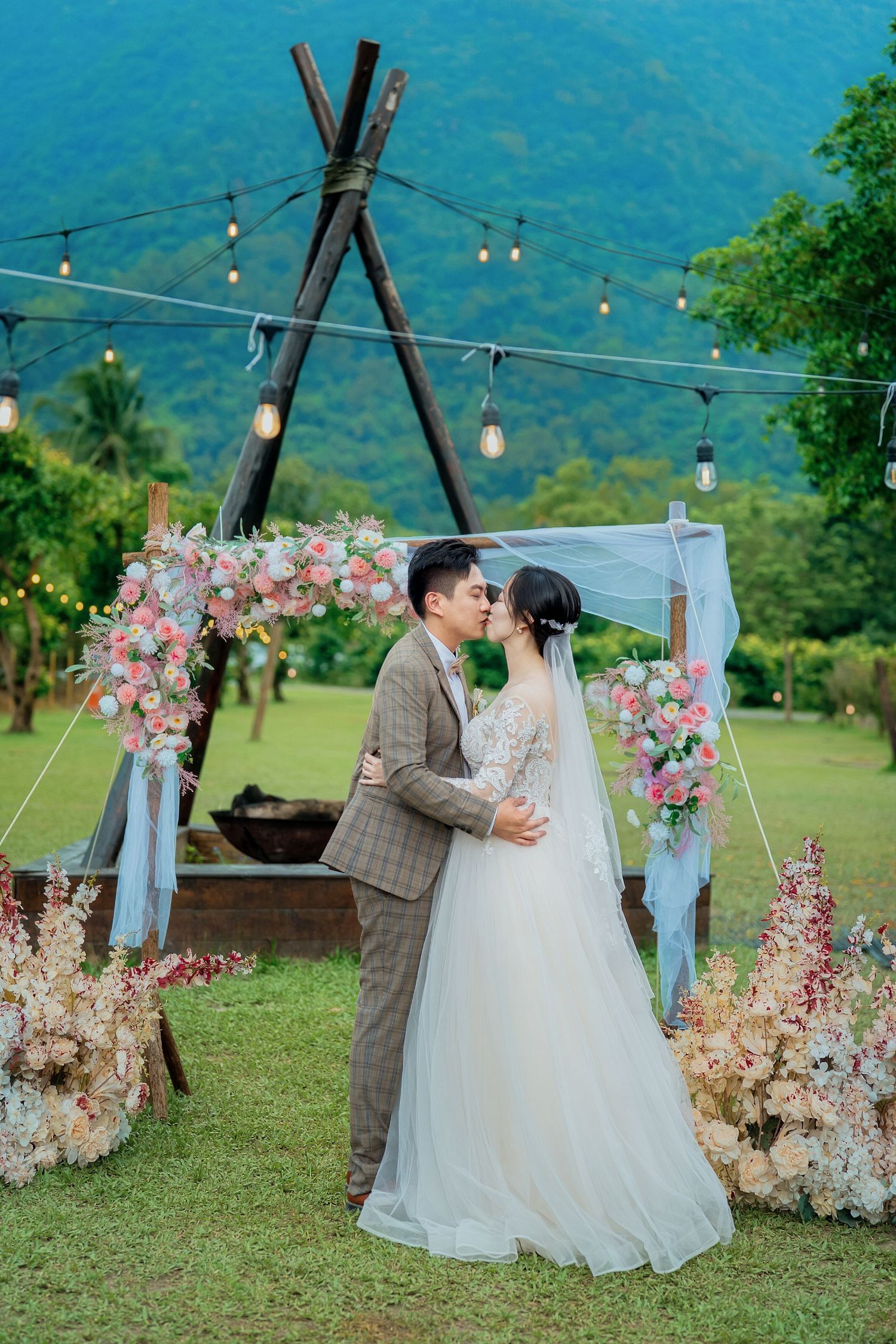 Romantic outdoor wedding ceremony with floral arch decorations, string lights, and mountain backdrop in grassy meadow.