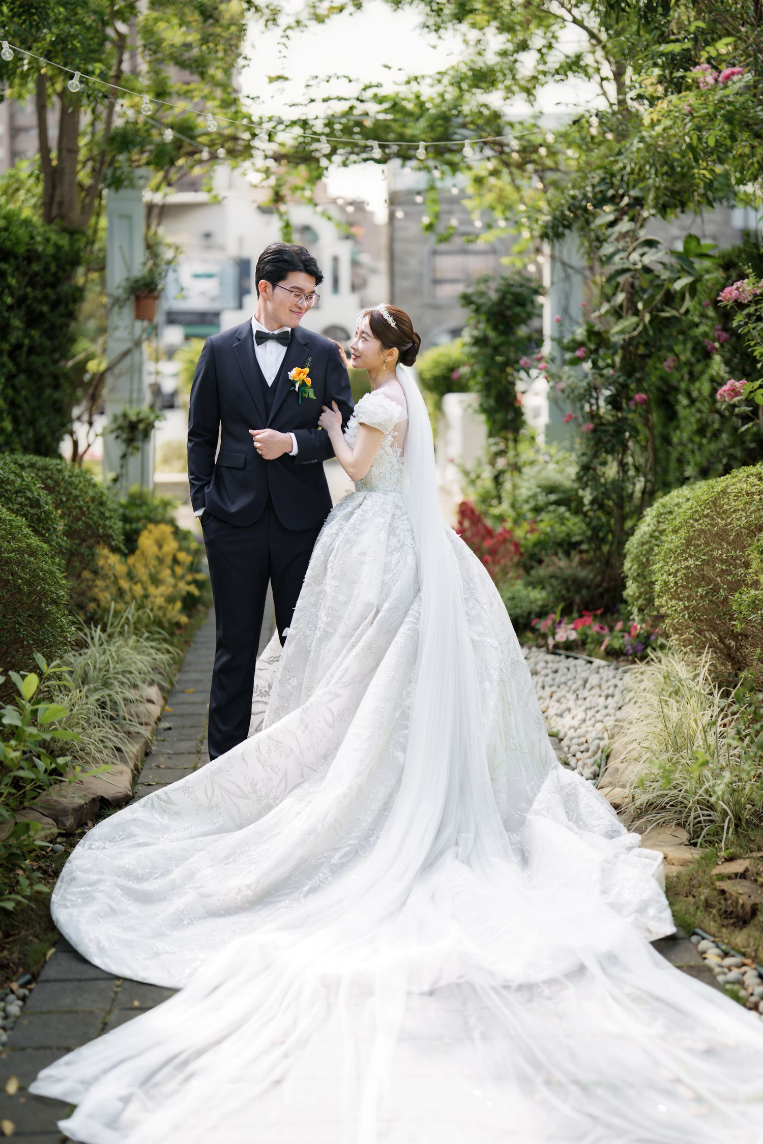 A couple in formal wedding attire poses together in a lush garden setting with a dramatic long white train on the dress.